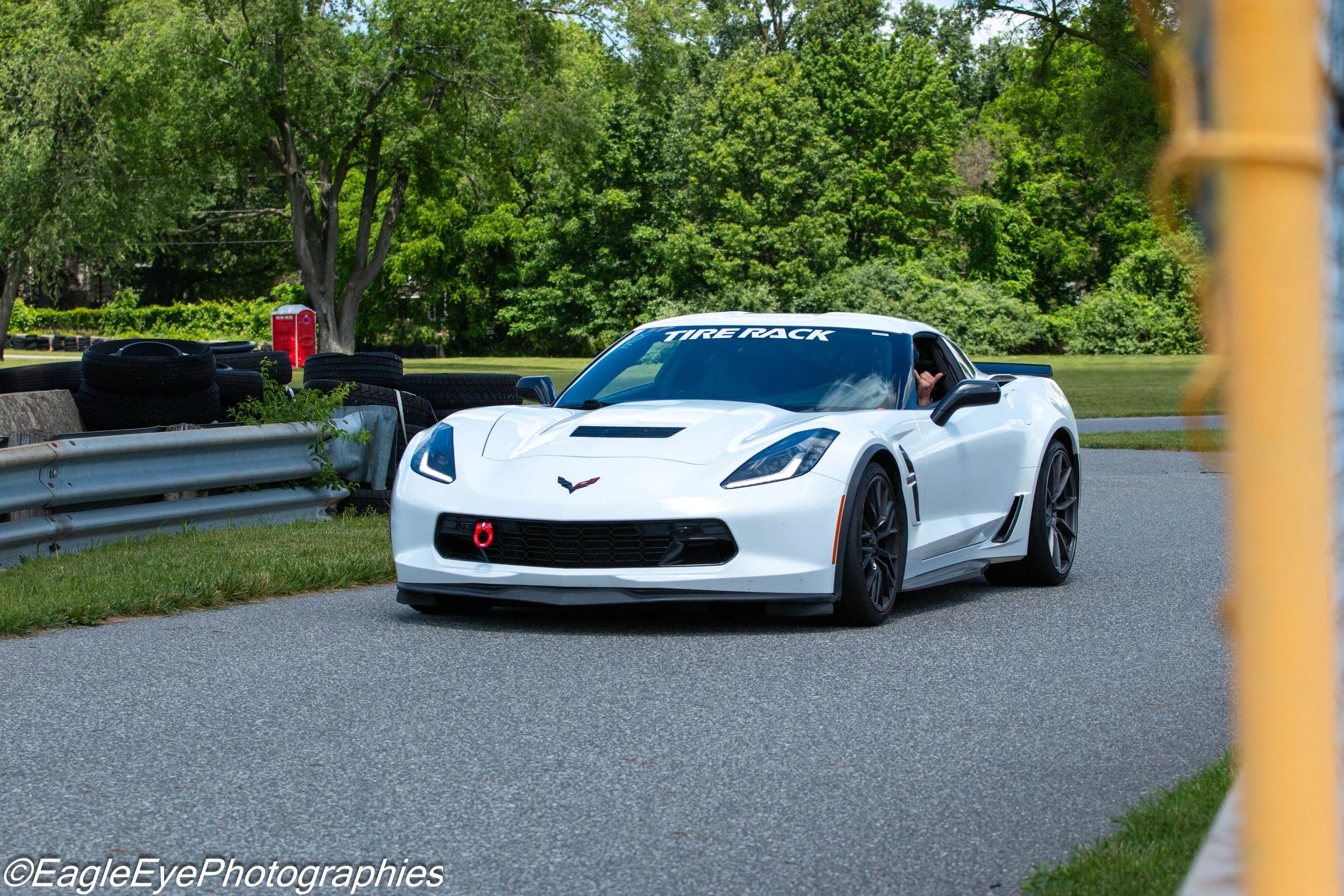 A white sports car with a black roof and #88 on the windshield, parked next to a stack of tires and a metal guardrail, with lush green trees and a red portable toilet in the background.
