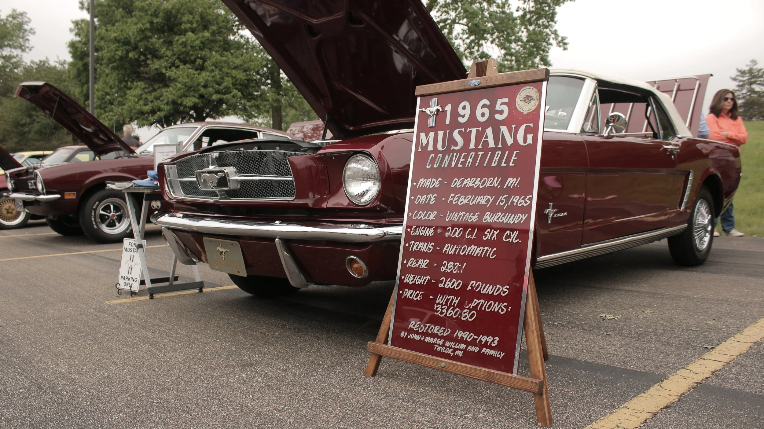 A 1965 red Ford Mustang convertible displayed at a car show, with a signboard providing details about the vehicle.