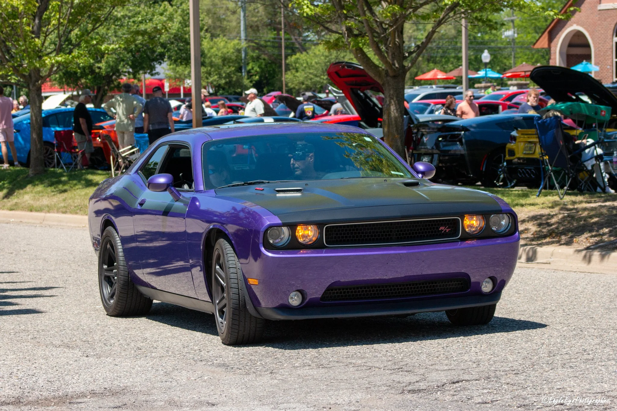 A purple Dodge Challenger sports car parked on a street during a car show with a crowd of people and other cars in the background.
