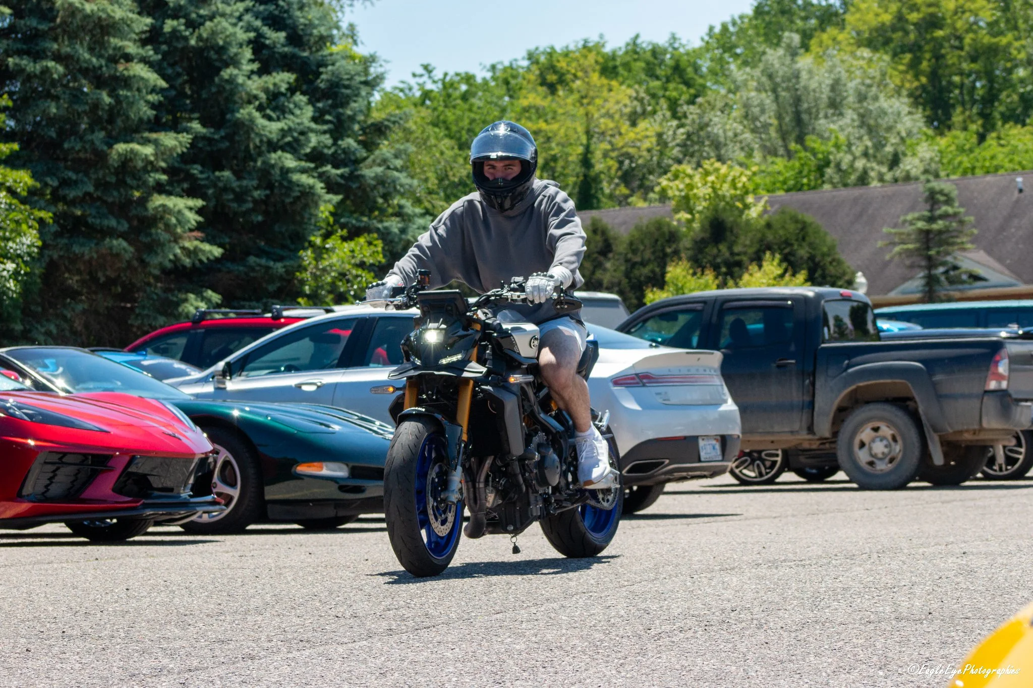 A person wearing a black helmet and gray hoodie riding a black motorcycle in a parking lot filled with cars surrounded by green trees.