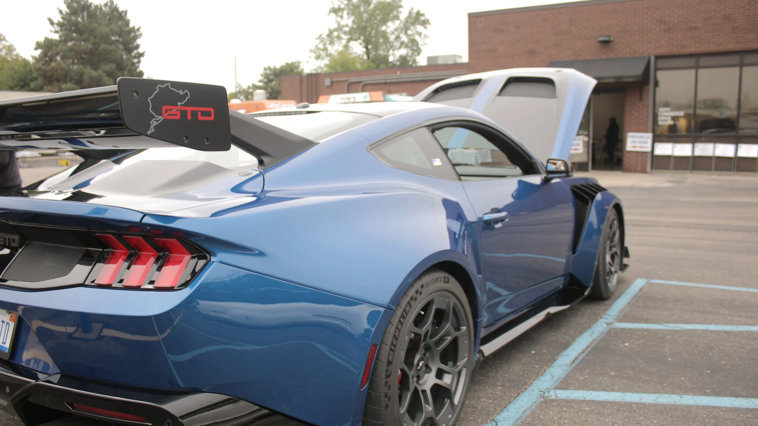 A blue sports car parked in a parking lot with the rear hood open, showing a large rear wing with a red sticker that reads 'GTD' and an outline of a race track.