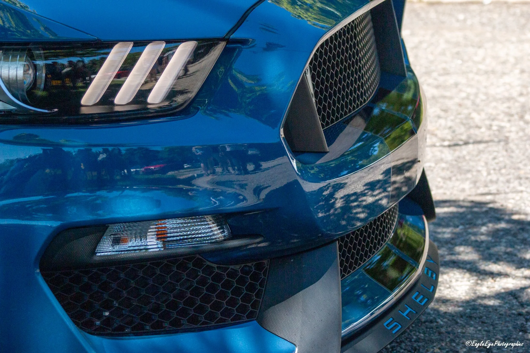 Close-up of a blue sports car's front, showing a headlight, grille, and a tire with the word 'SHIELD' visible.