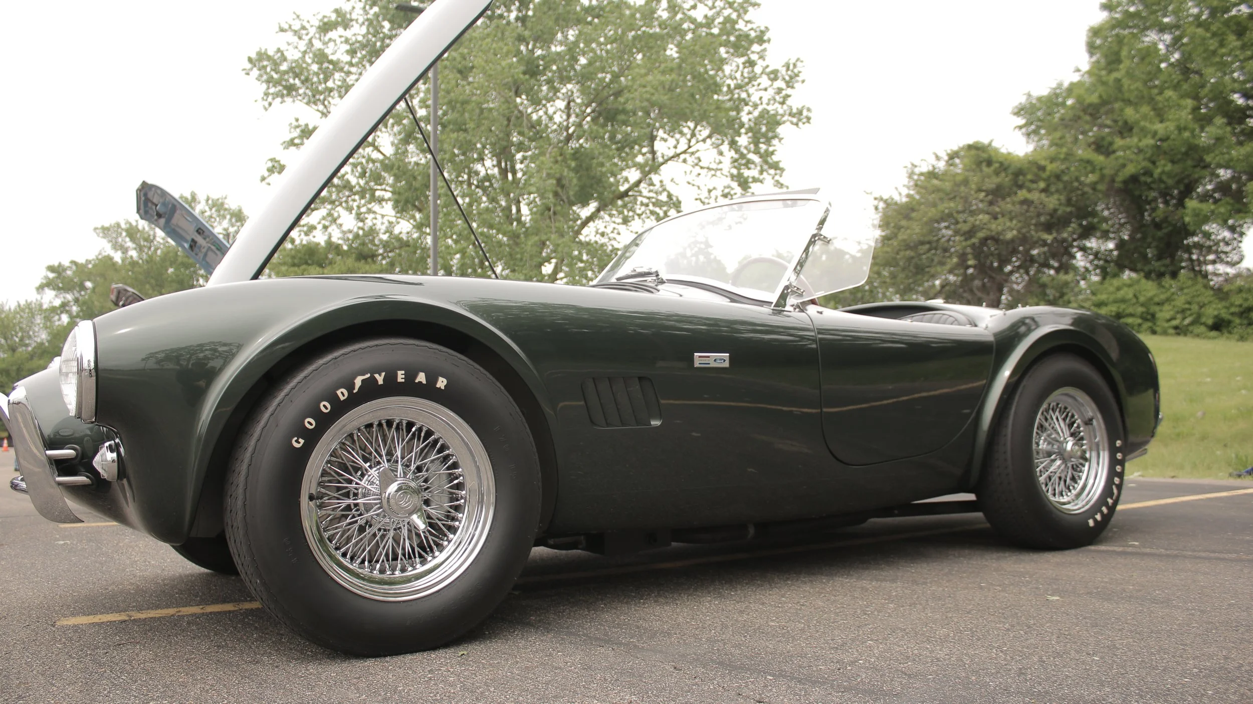 A classic black convertible sports car with chrome wire wheels and tire branding, parked outdoors on a paved lot with green trees in the background and a partially open hood.