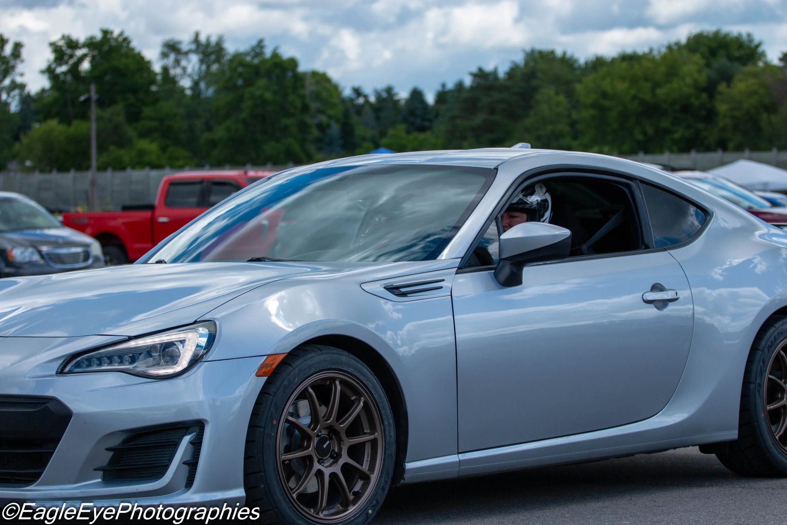 A silver sports car with a driver wearing a racing helmet, parked among other vehicles at an outdoor event with trees and cloudy skies in the background.