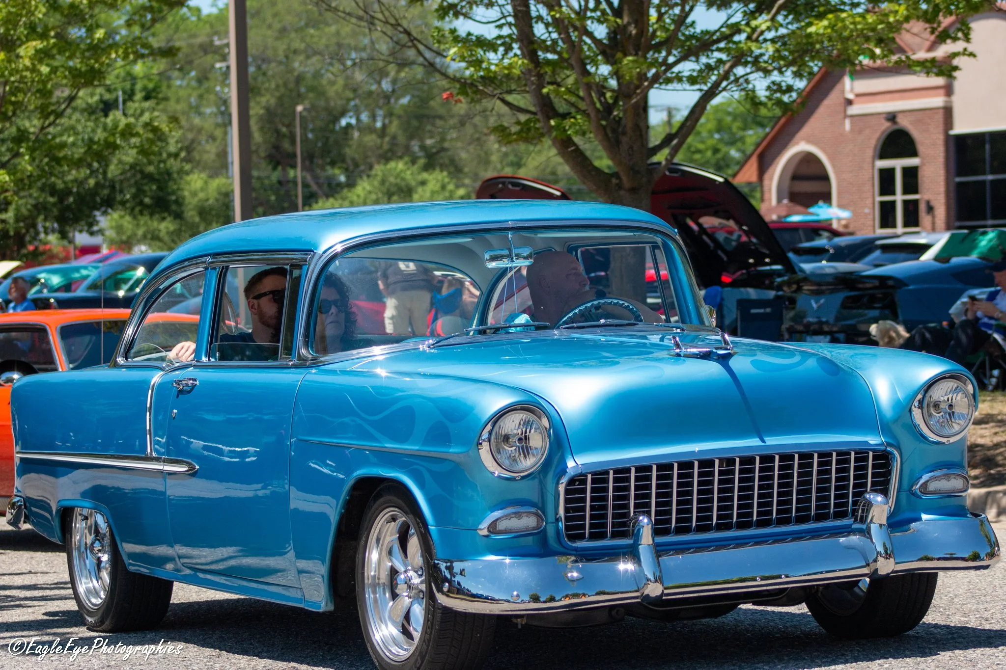A blue classic vintage car at an outdoor event, surrounded by other vehicles and spectators, with trees and a brick building in the background.