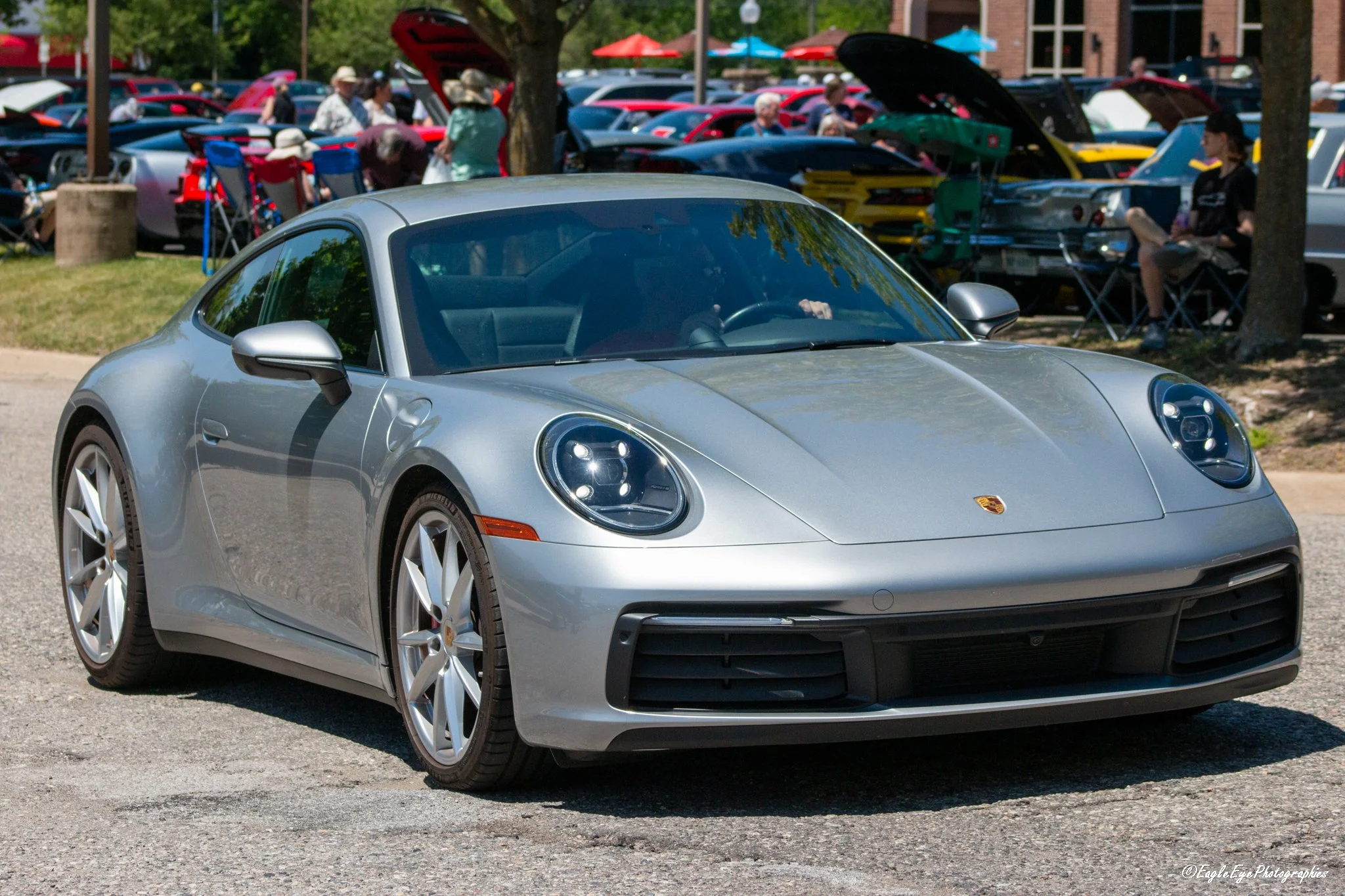 Silver Porsche sports car parked at a crowded outdoor car show with people and other vehicles in the background.