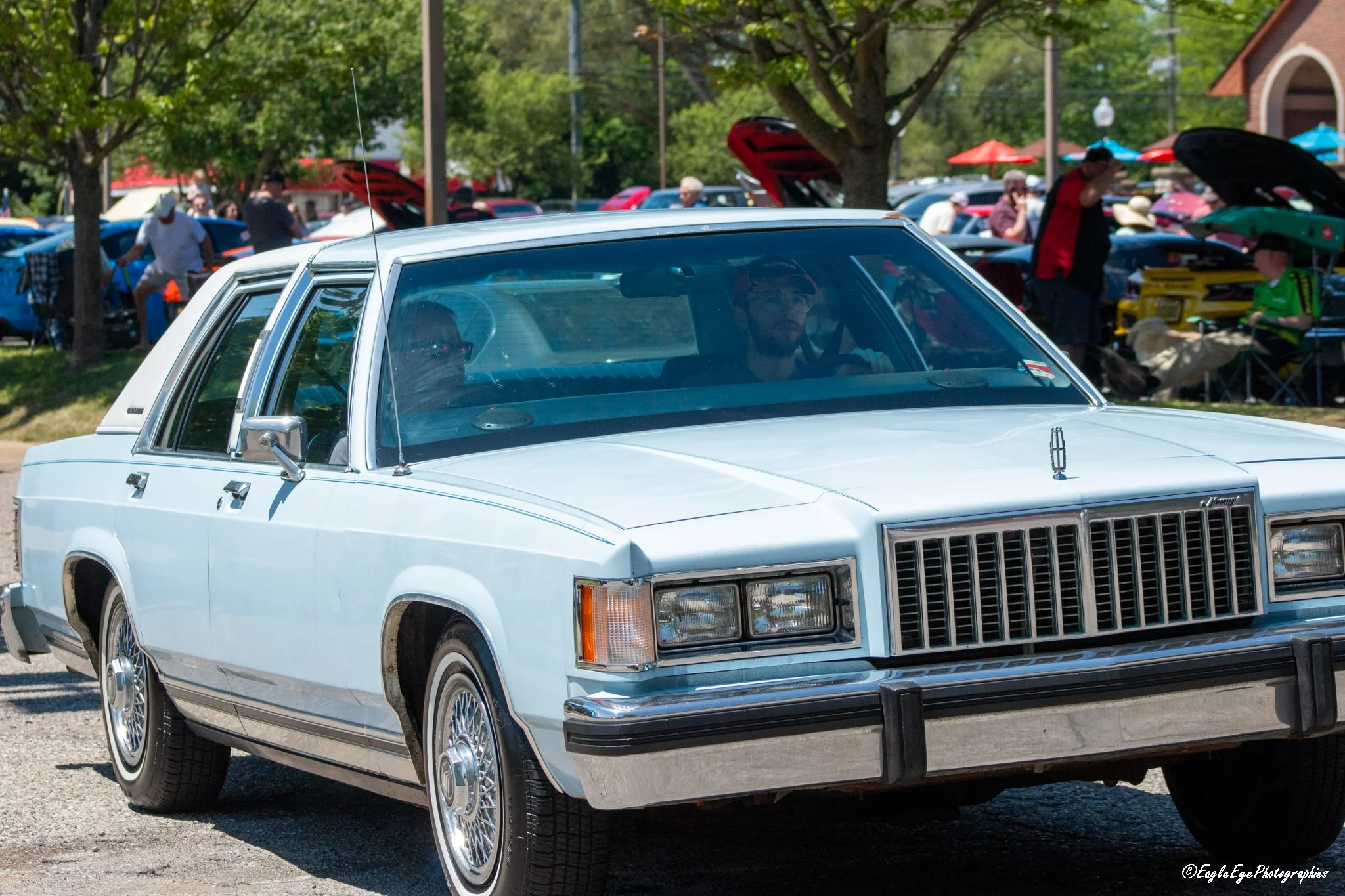 A vintage white car with chrome accents parked at an outdoor car show, with people walking around and other cars in the background.