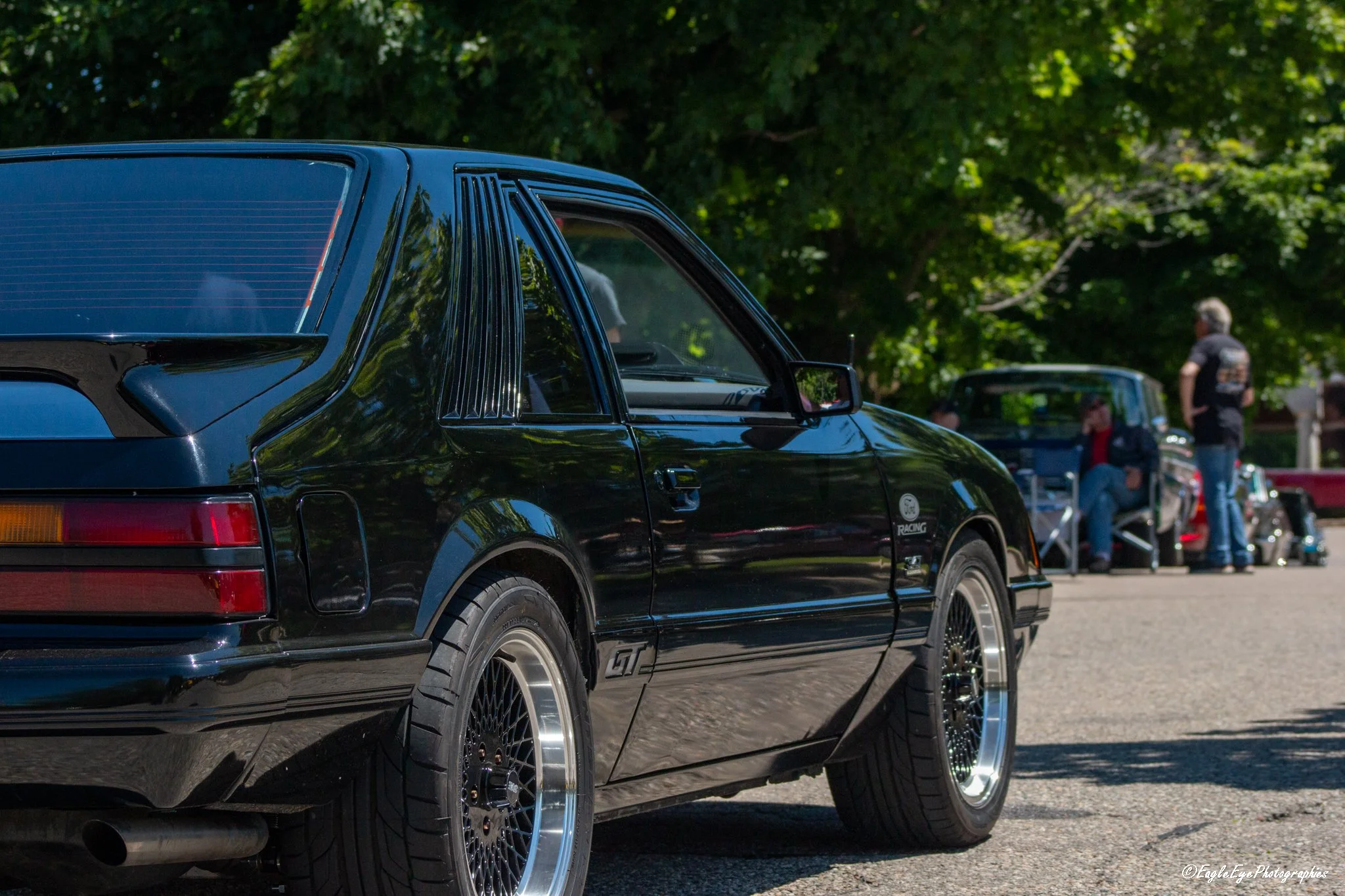 Close-up of a black vintage car with custom wheels parked outdoors during daytime, with trees and people in the background.