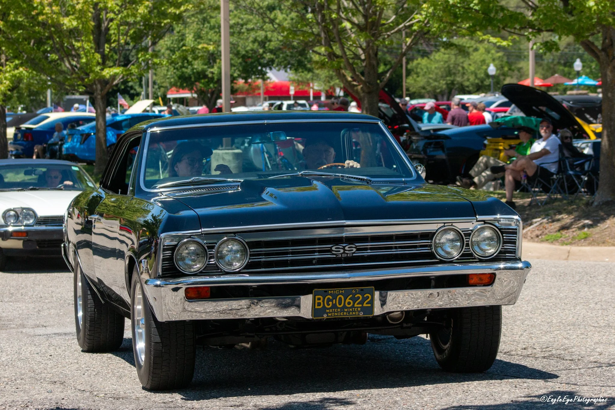 Black 1967 Chevrolet Chevelle SS 396 with Michigan license plate in a car show setting, surrounded by other classic cars and people under trees.