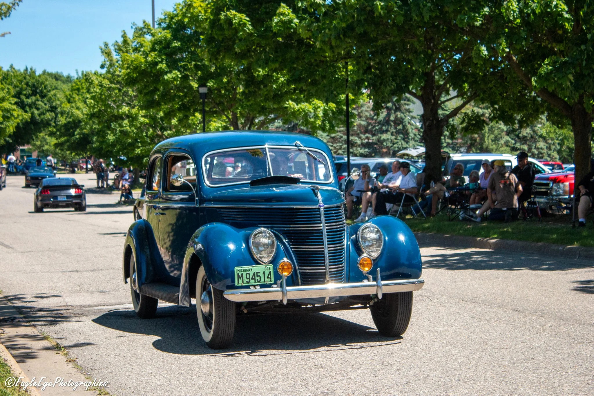 A vintage blue car with a Michigan license plate parked in a park surrounded by people sitting on benches and enjoying a sunny day under green trees.