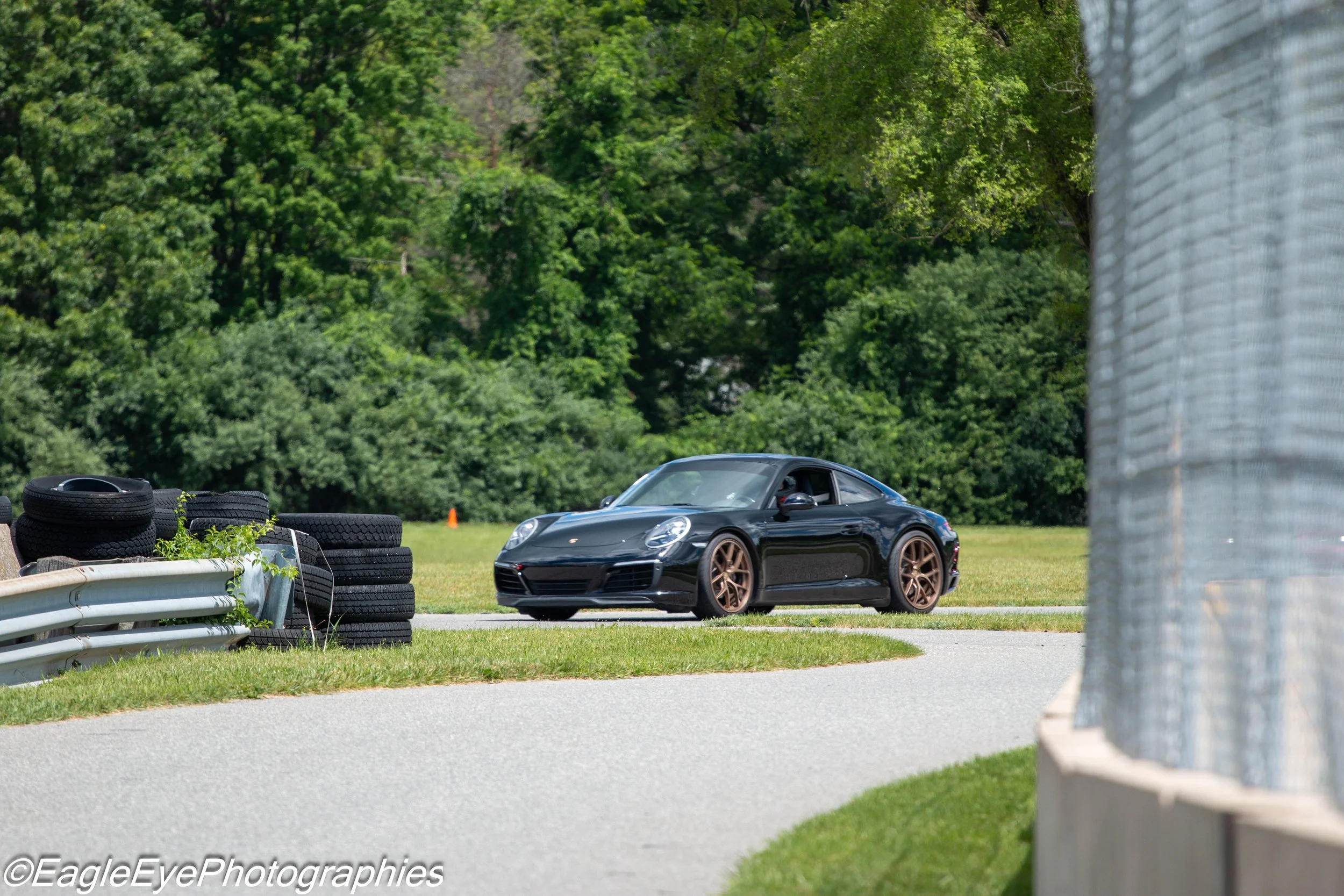 A black sports car parked on a grassy area next to a stack of tires and a guardrail, with a background of dense green trees.