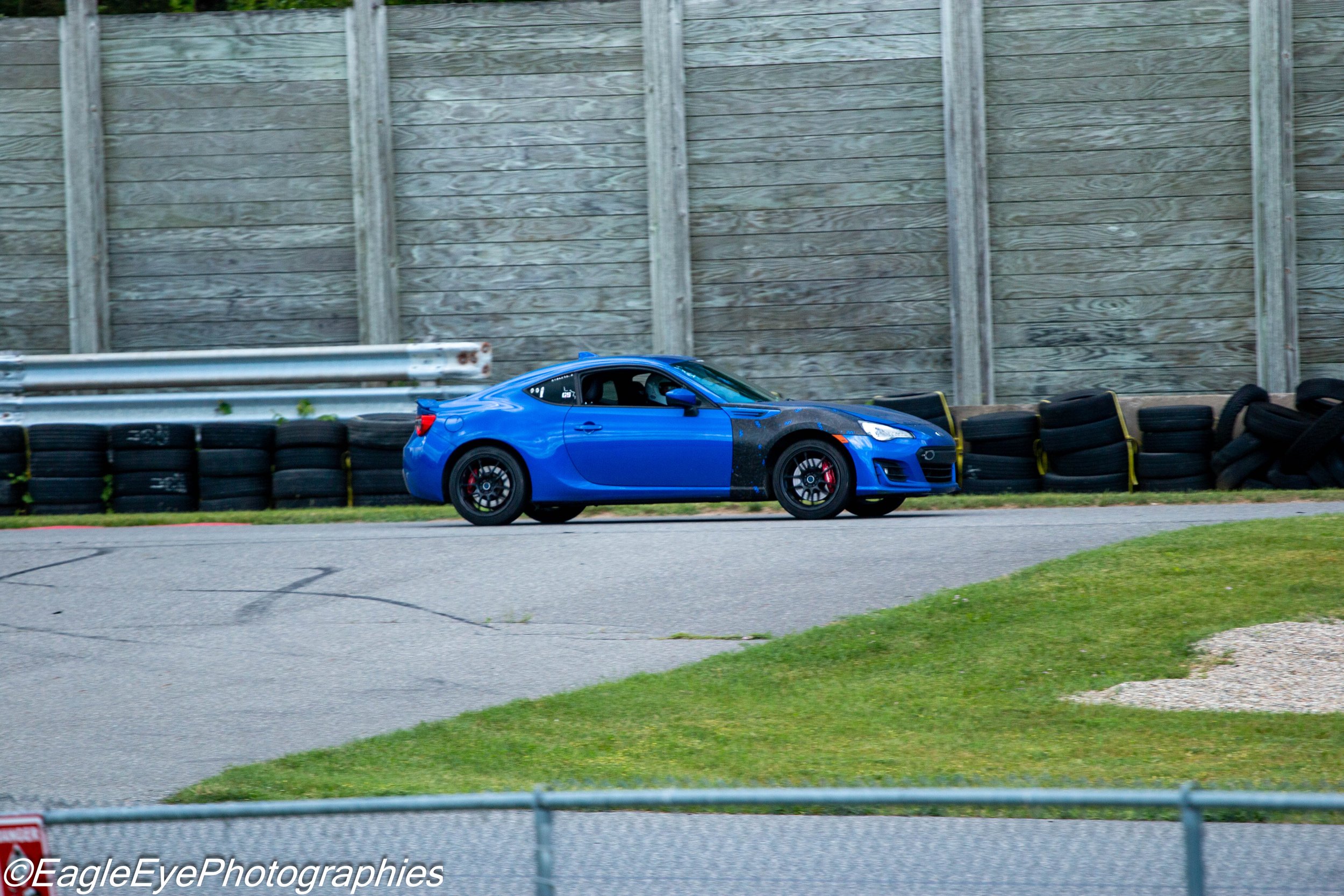 A blue sports car driving on a racetrack, with a wooden barrier and tires on the side.