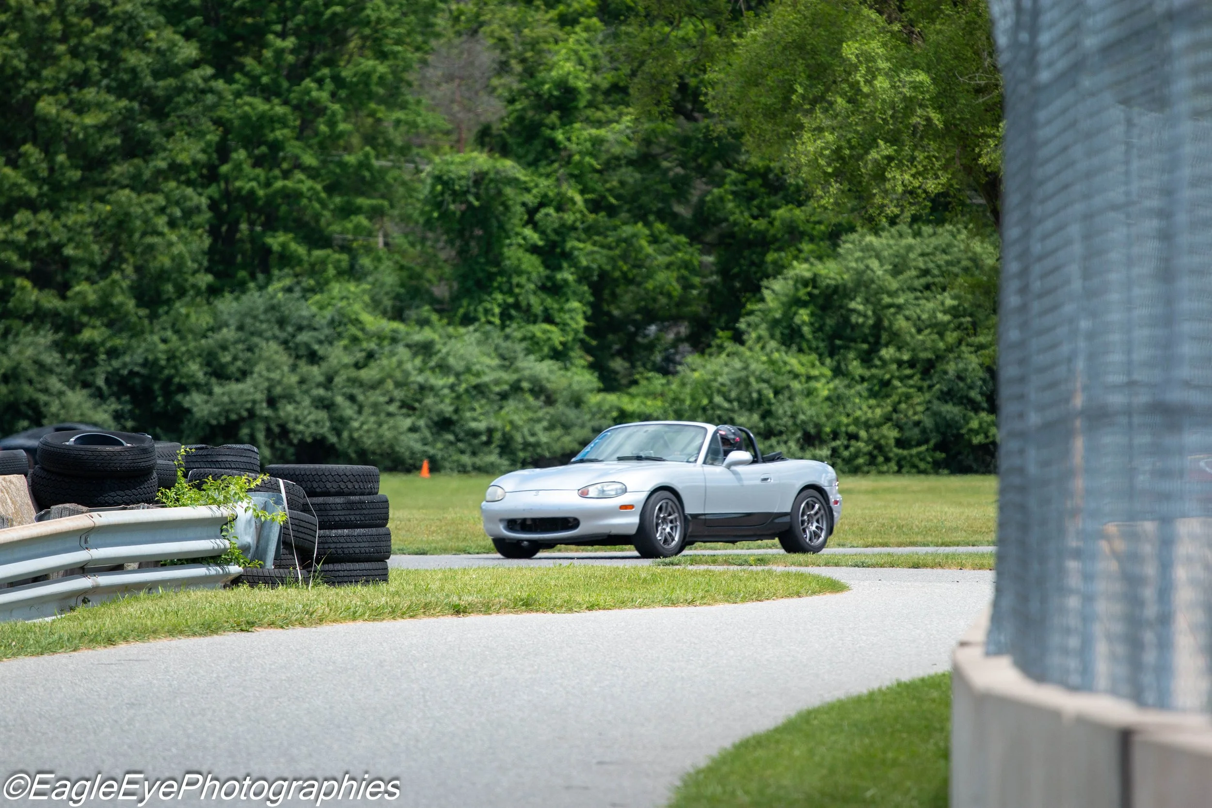 A white convertible car parked on a grassy area near a tire barrier at a racing track, with a background of dense green trees.
