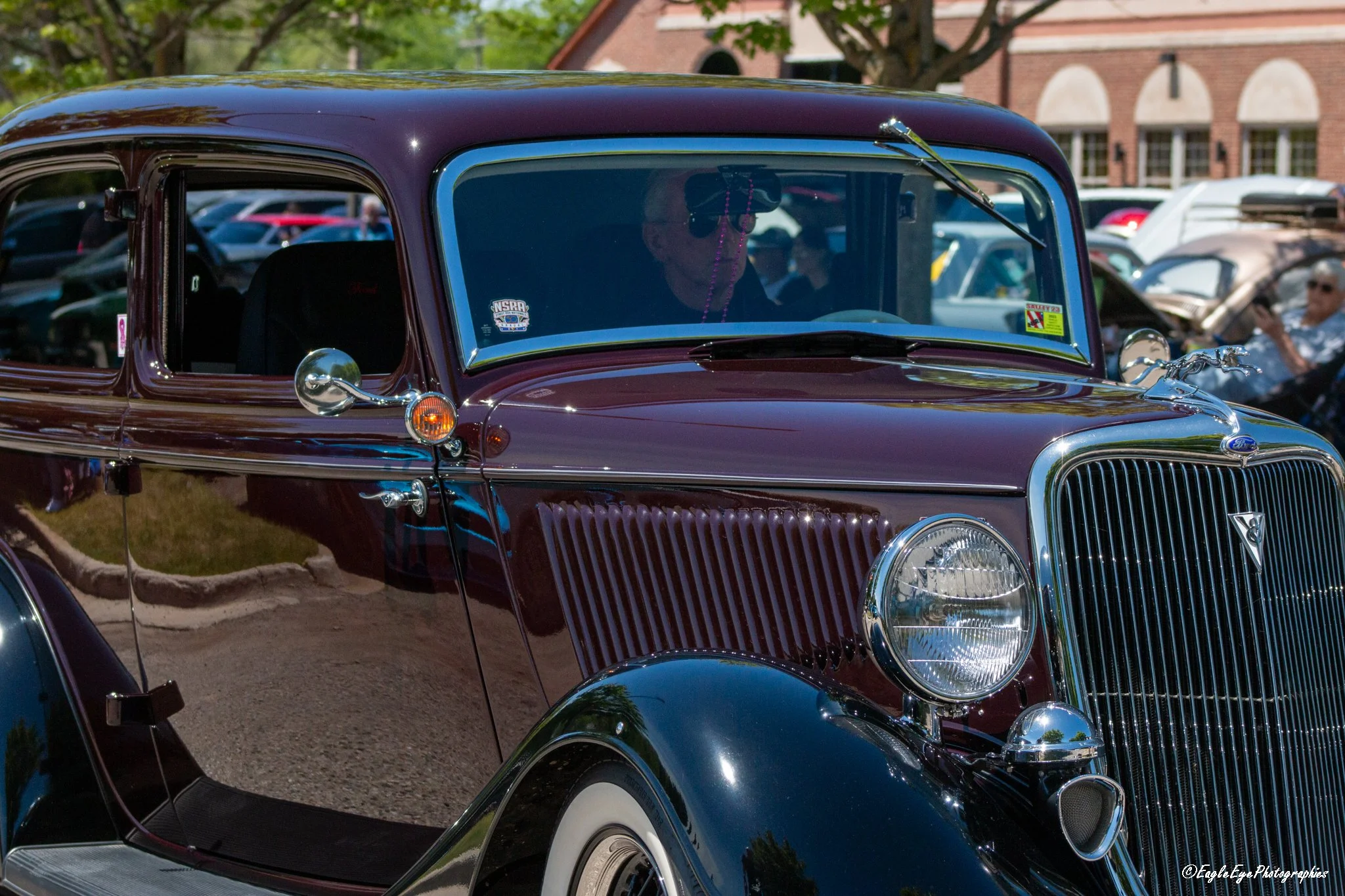 A vintage maroon and black classic car with chrome details on display outdoors at a car show, with trees and a brick building in the background.