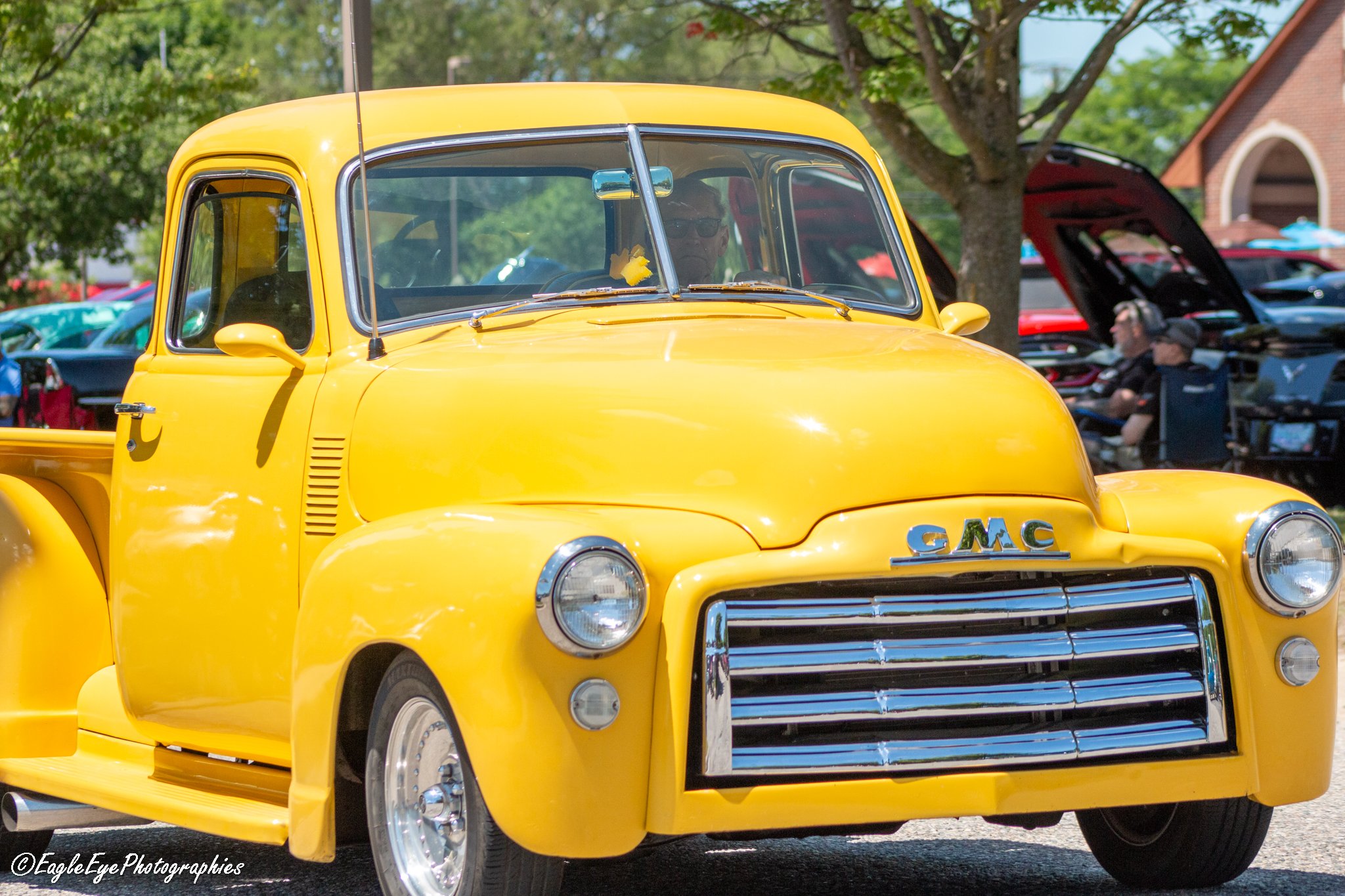 A bright yellow vintage GMC pickup truck on display at an outdoor car show, with a person visible through the windshield and other classic cars and trees in the background.