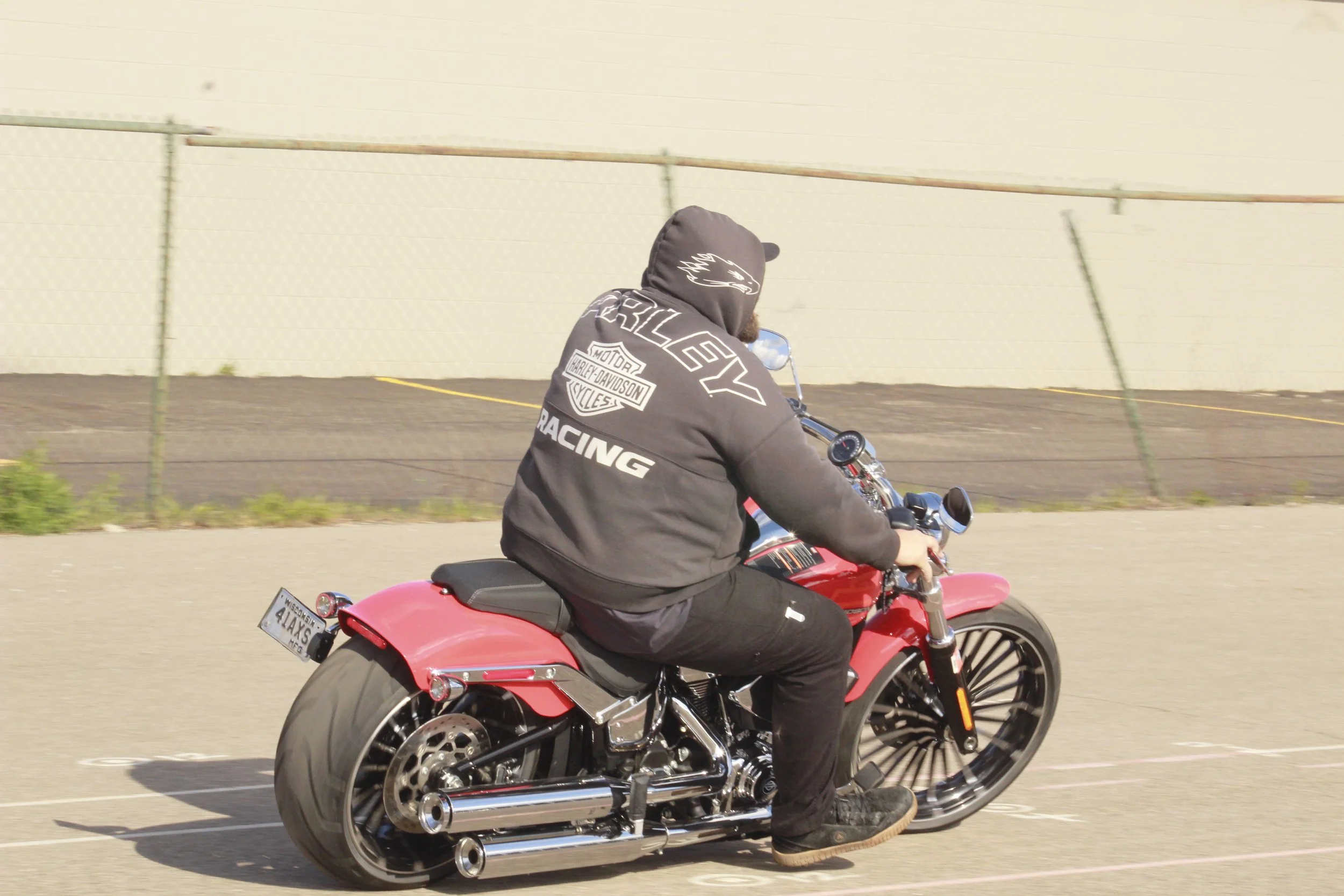 Person wearing a Harley-Davidson hoodie riding a red motorcycle on the street with a chain link fence and a light-colored building in the background.