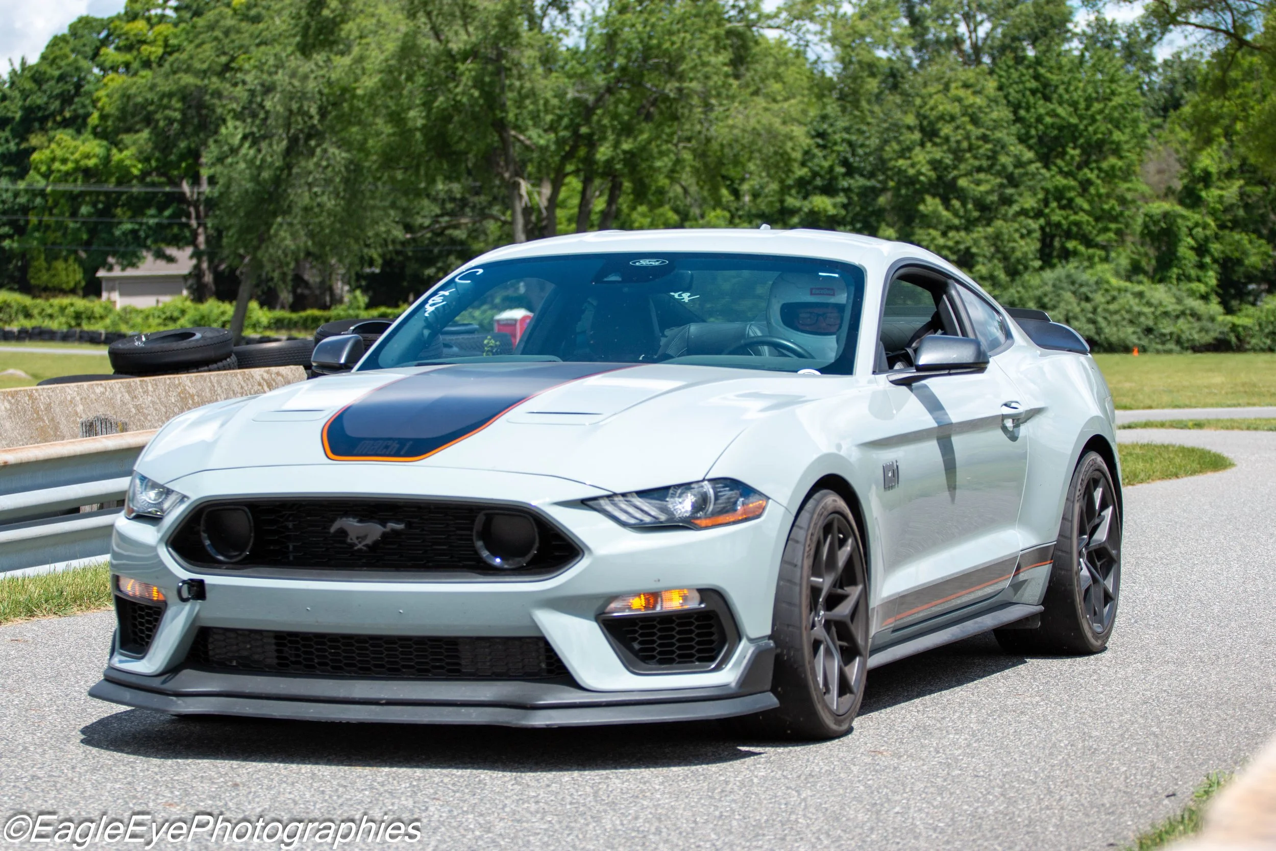 A white Ford Mustang sports car with black racing stripes on the hood, black wheels, and a black spoiler, driving on a road with trees and greenery in the background.
