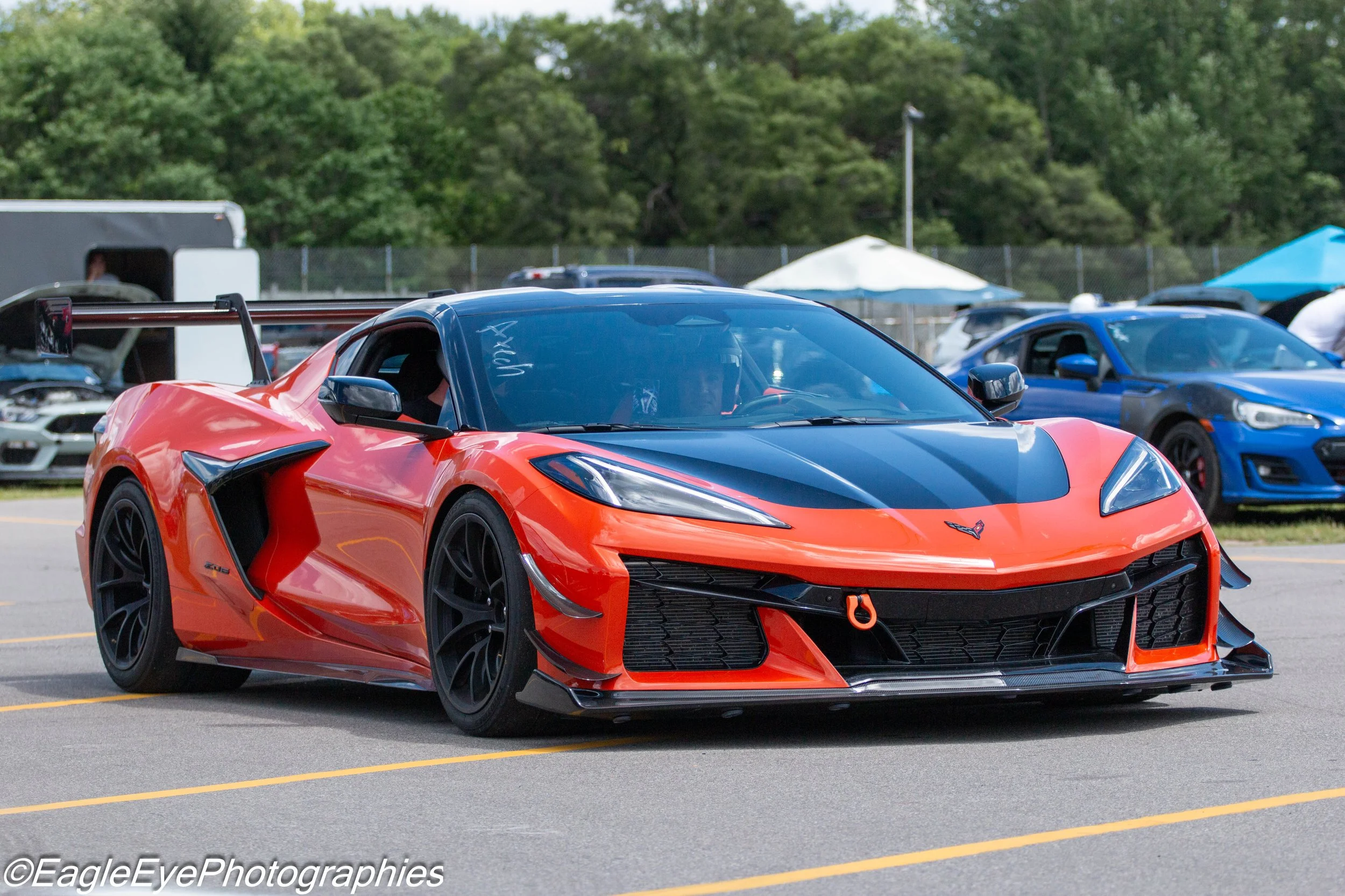 A red and black Chevrolet Corvette sports car with a racing wing is parked in a parking lot during a car event, with other cars, tents, and trees in the background.