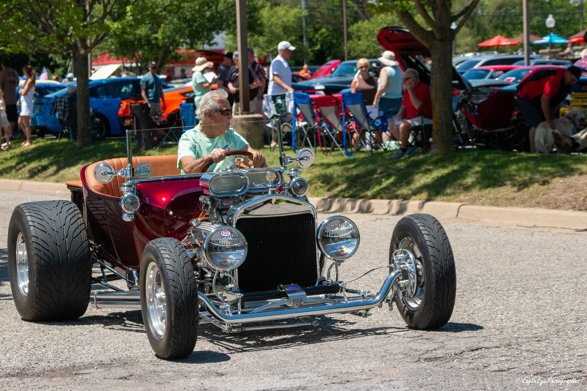 A vintage-style small car with large tires being driven at a car show with classic and modern cars in the background and spectators on the grass and under umbrellas.