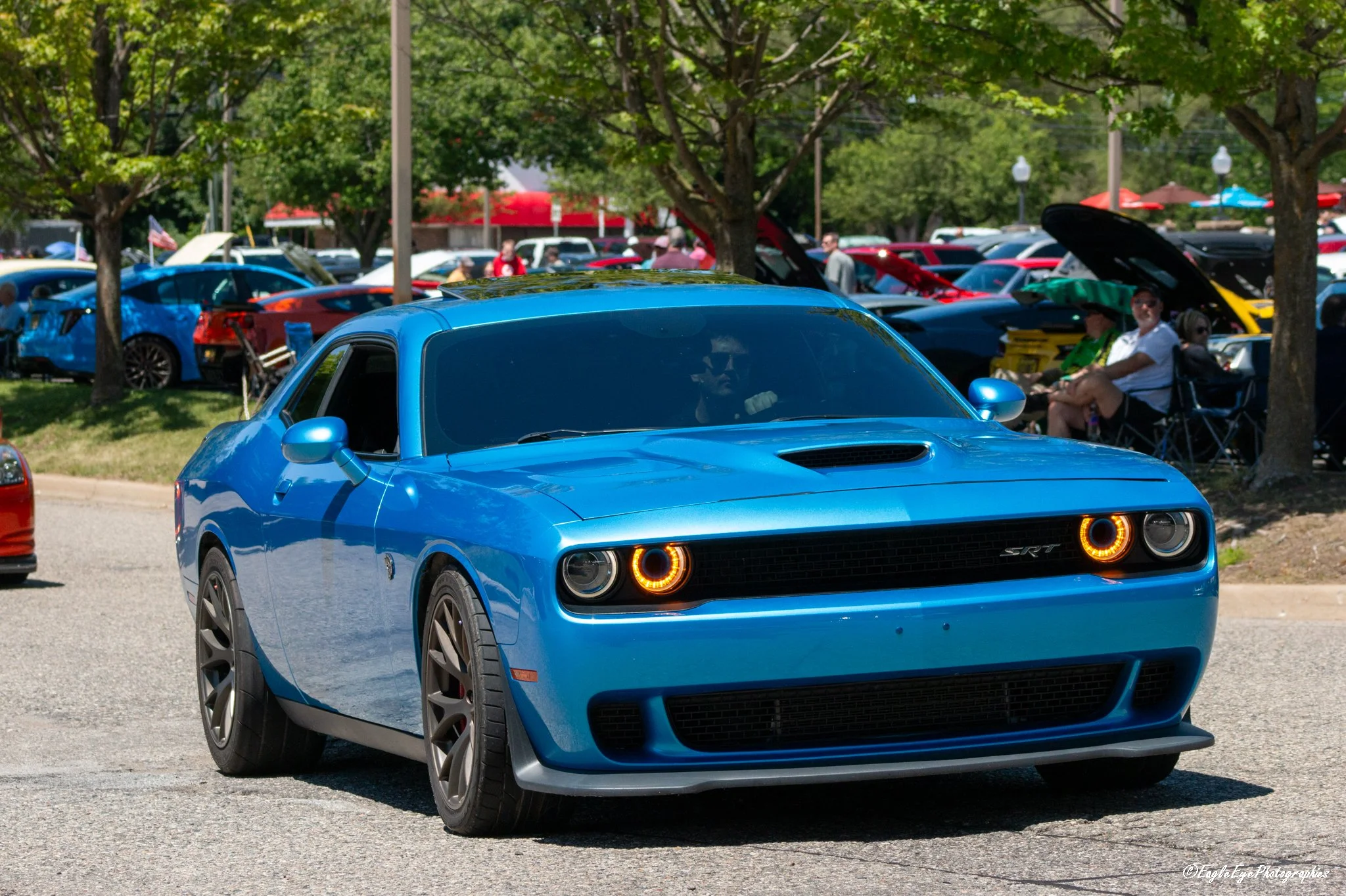A blue muscle car driving on a street during an outdoor event, with people sitting and walking in the background among trees and parked cars.