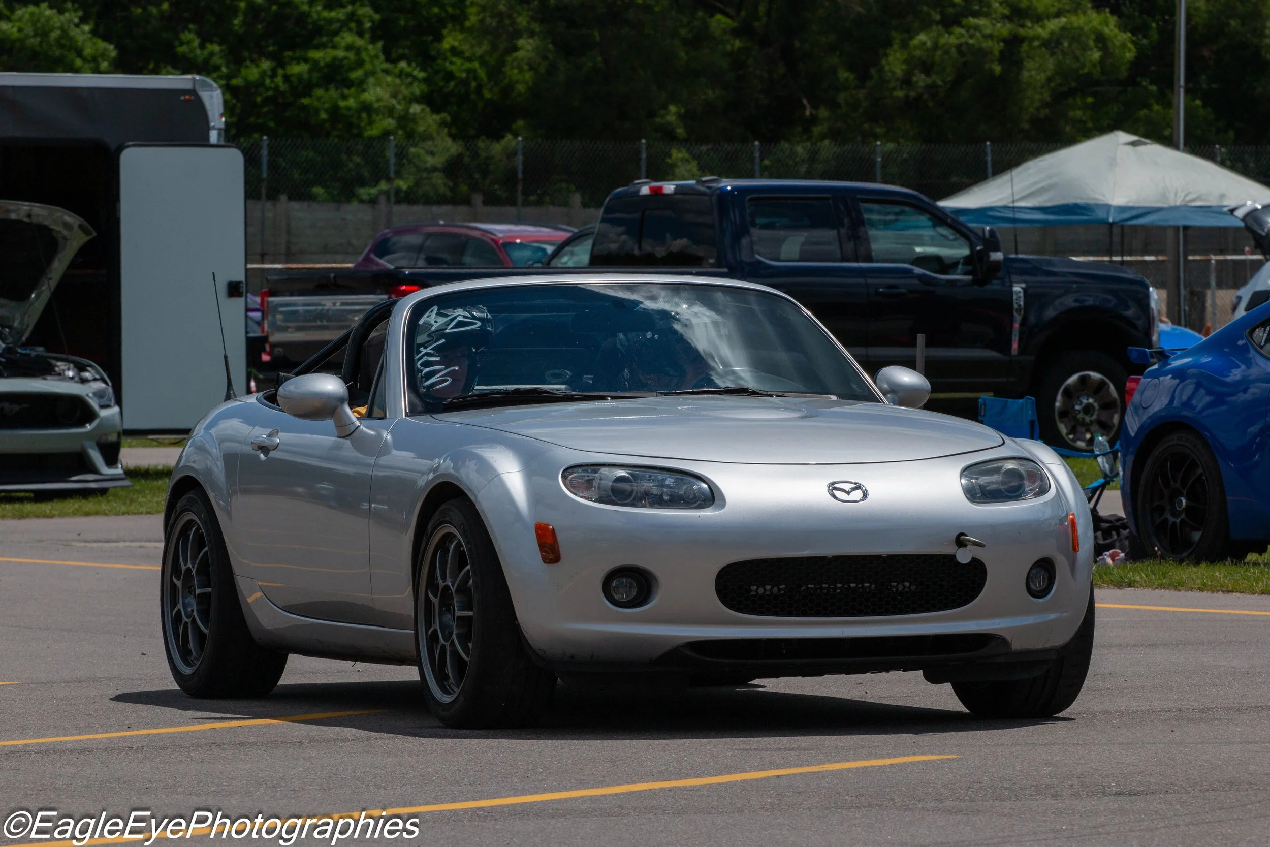 A silver Mazda MX-5 Miata convertible parked in a lot with other vehicles, including a black pickup truck, behind a fencing and trees, with a white tent visible in the background.