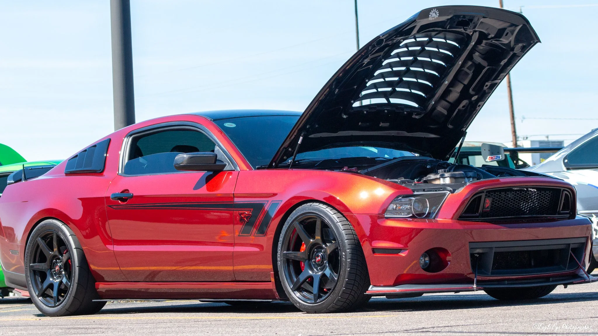 Red Ford Mustang sports car with open hood parked outdoors, showing engine details, alongside other cars at a car show.
