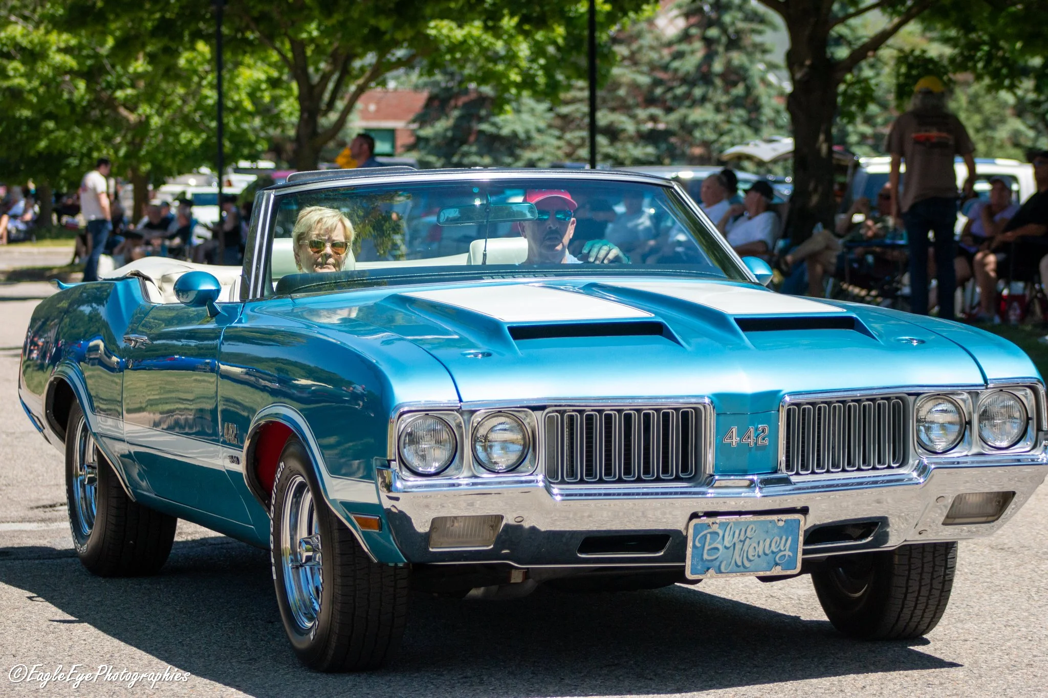 A vintage blue convertible car with a white interior, driven by a man wearing sunglasses and a red cap, with a woman in the passenger seat. The background shows a park with people sitting on benches and standing near trees, under a sunny sky.