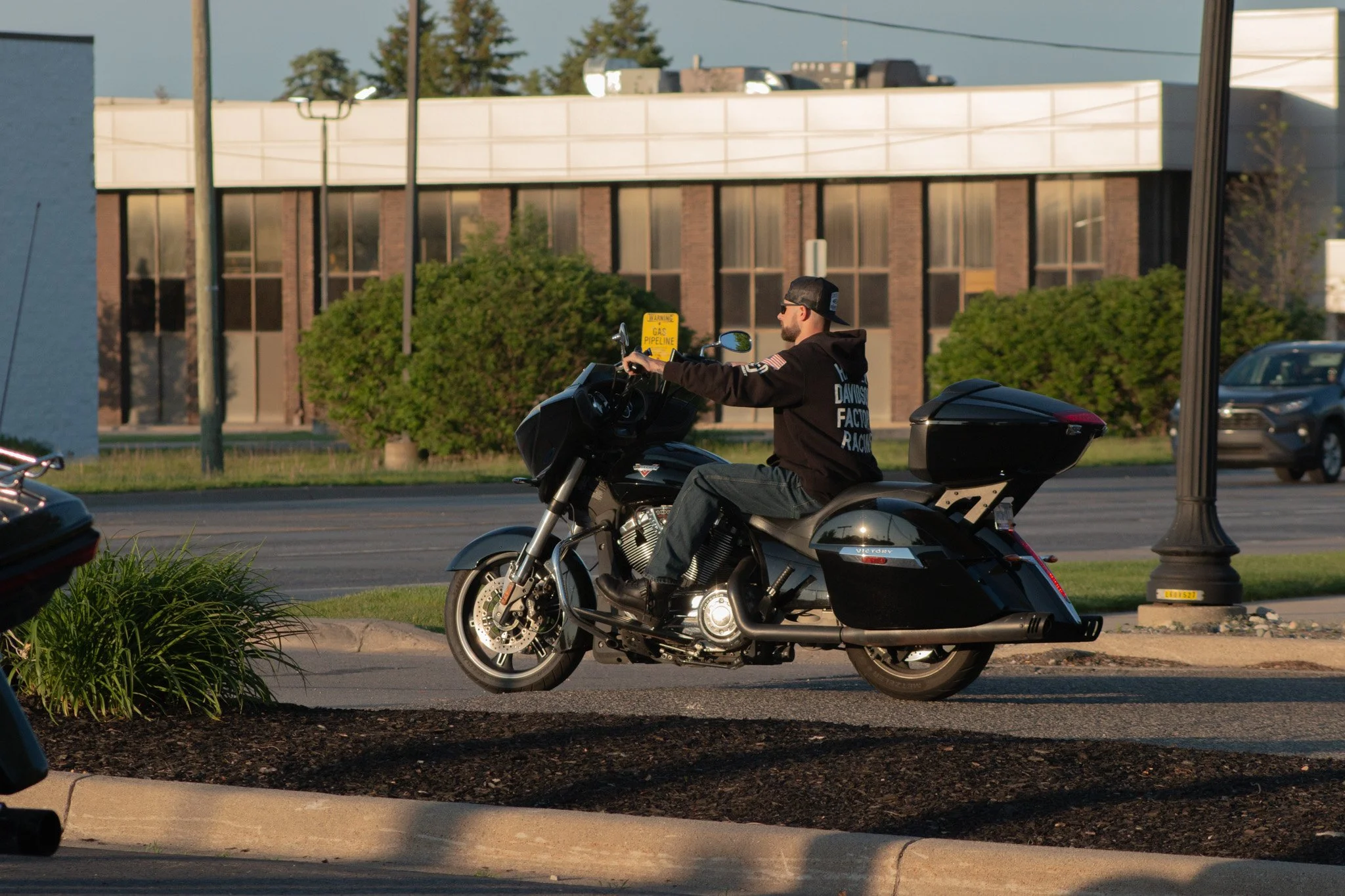 A man riding a black Harley-Davidson motorcycle on a city street, wearing a black hoodie with text on the back and a backwards cap, with trees, a building, and cars in the background.