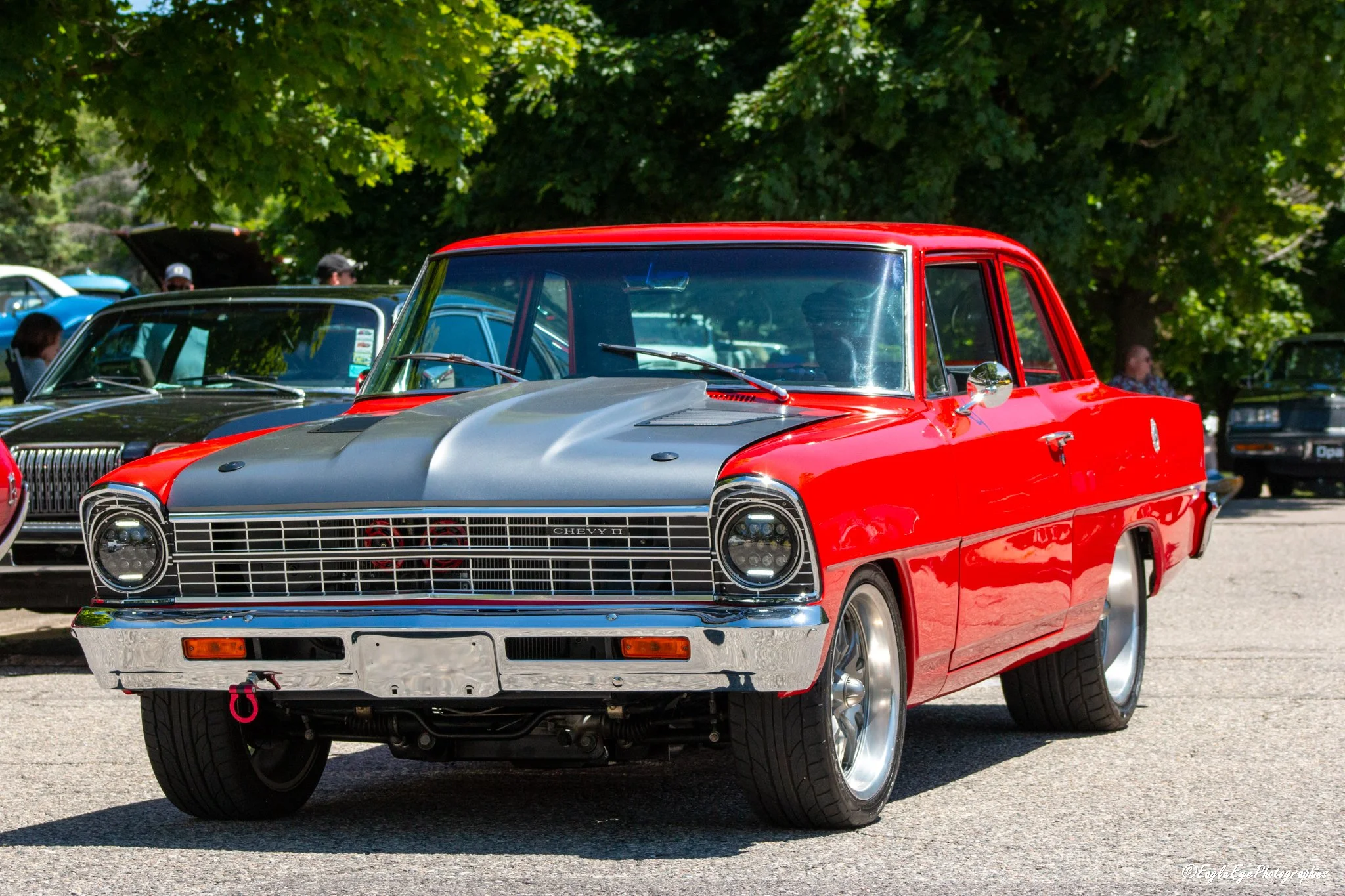 A red and black vintage Chevrolet car on display at a car show, with several other classic vehicles and people in the background under large trees.
