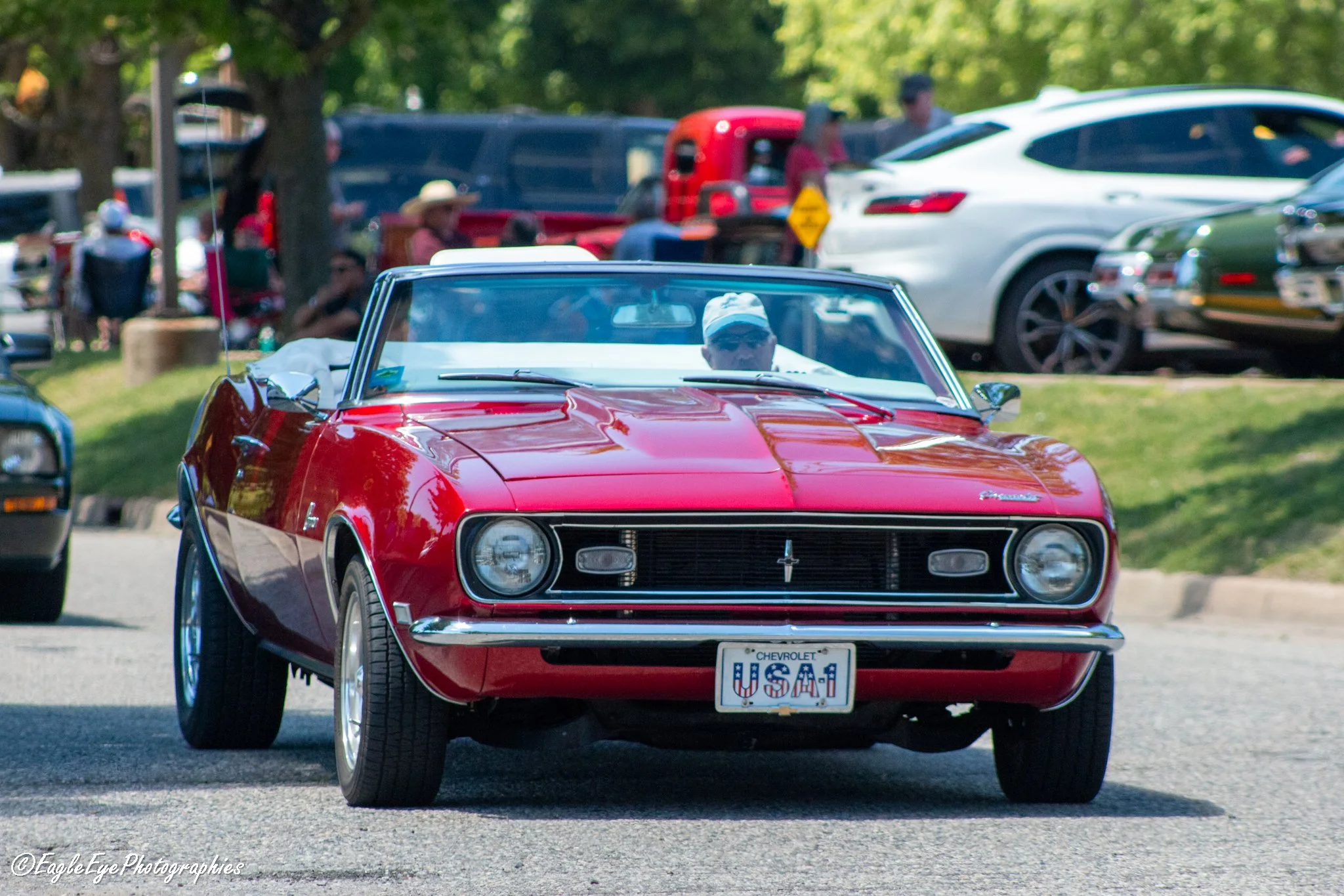 Red vintage Chevrolet Corvette driving on a street, with several parked cars and green trees in the background.