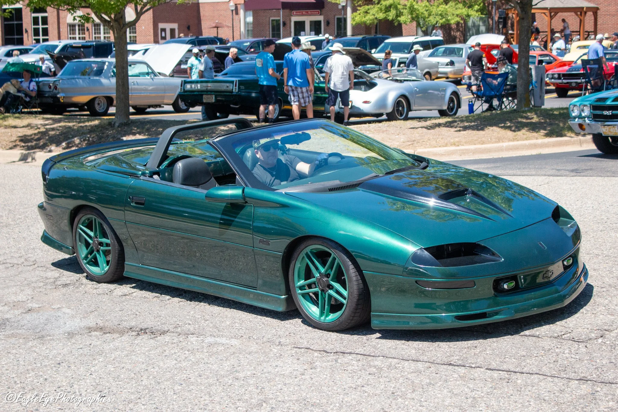 A green Chevrolet Corvette convertible parked at a car show during the daytime on a sunny day, with people walking around and looking at various vintage and classic cars in the background.