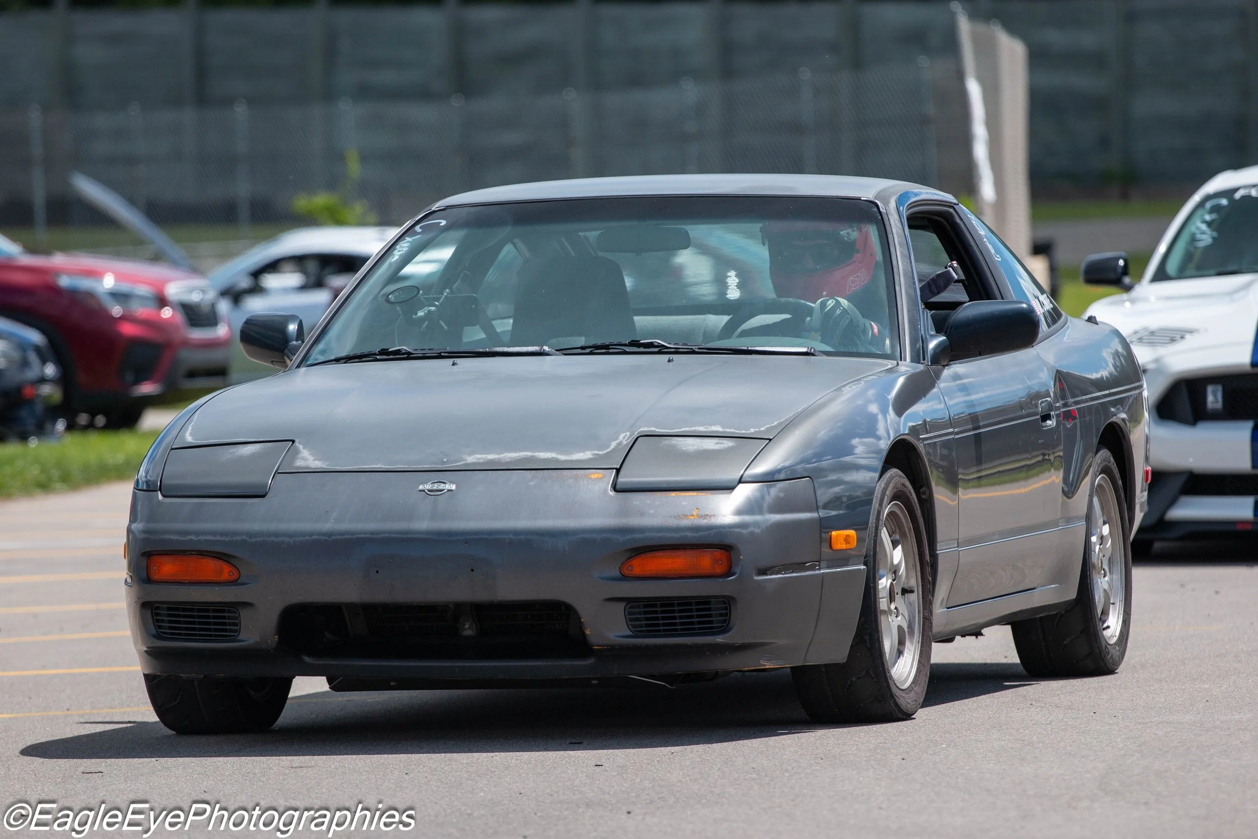 A silver Nissan 240SX sports car is driving on a parking lot, with other cars visible in the background.