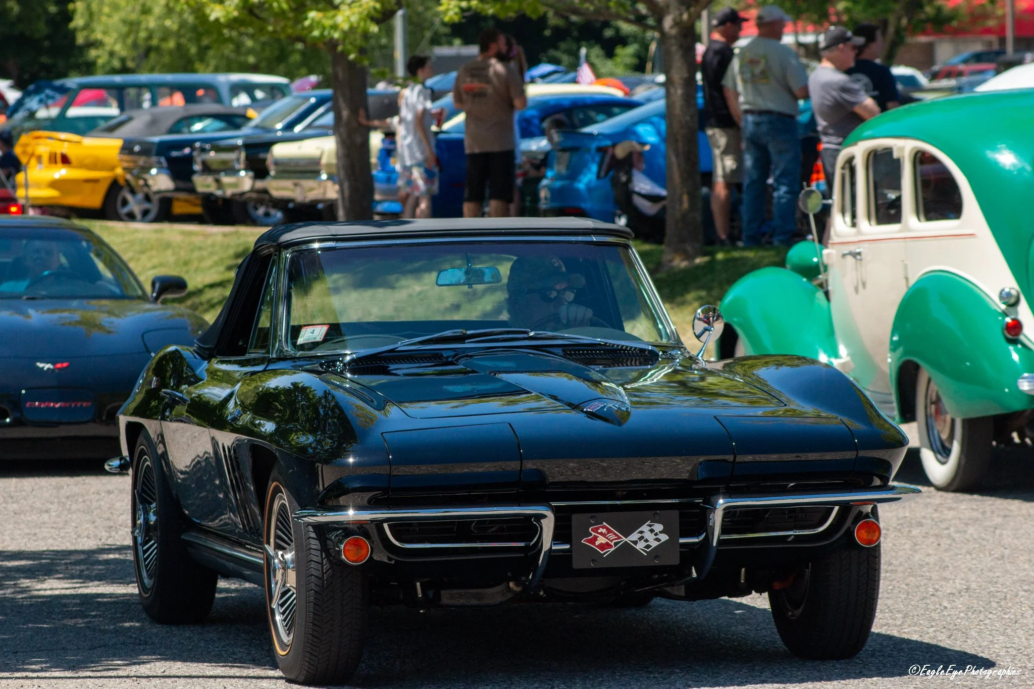 A vintage black Corvette sports car in front of a row of classic cars at a car show, with people walking and viewing the vehicles in the background.