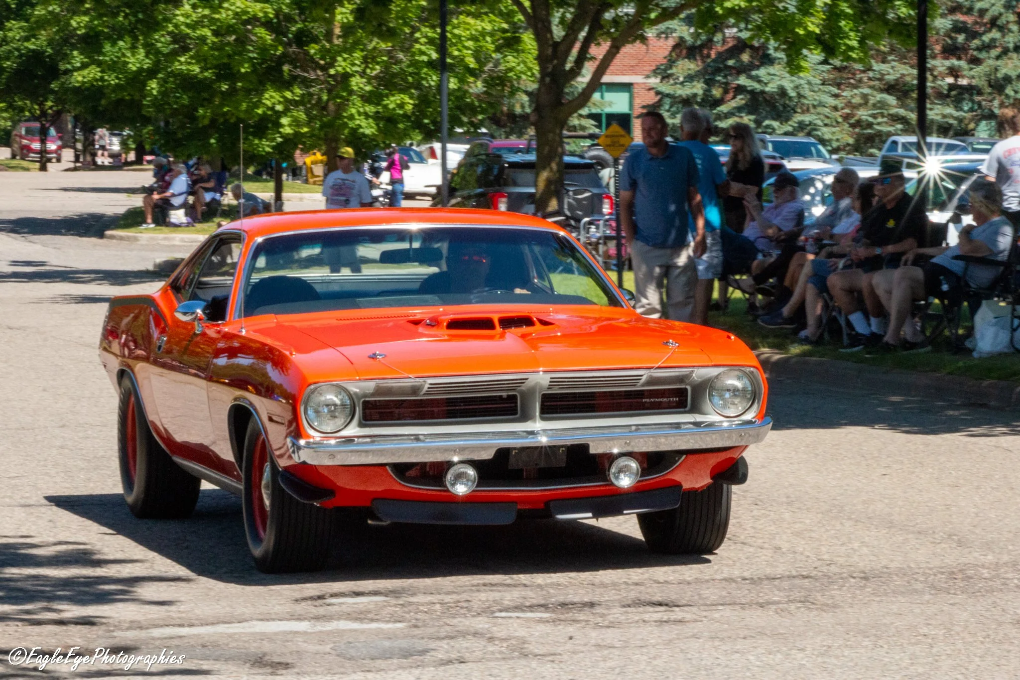 A red vintage muscle car driving down a street during a car show event with spectators and trees in the background.