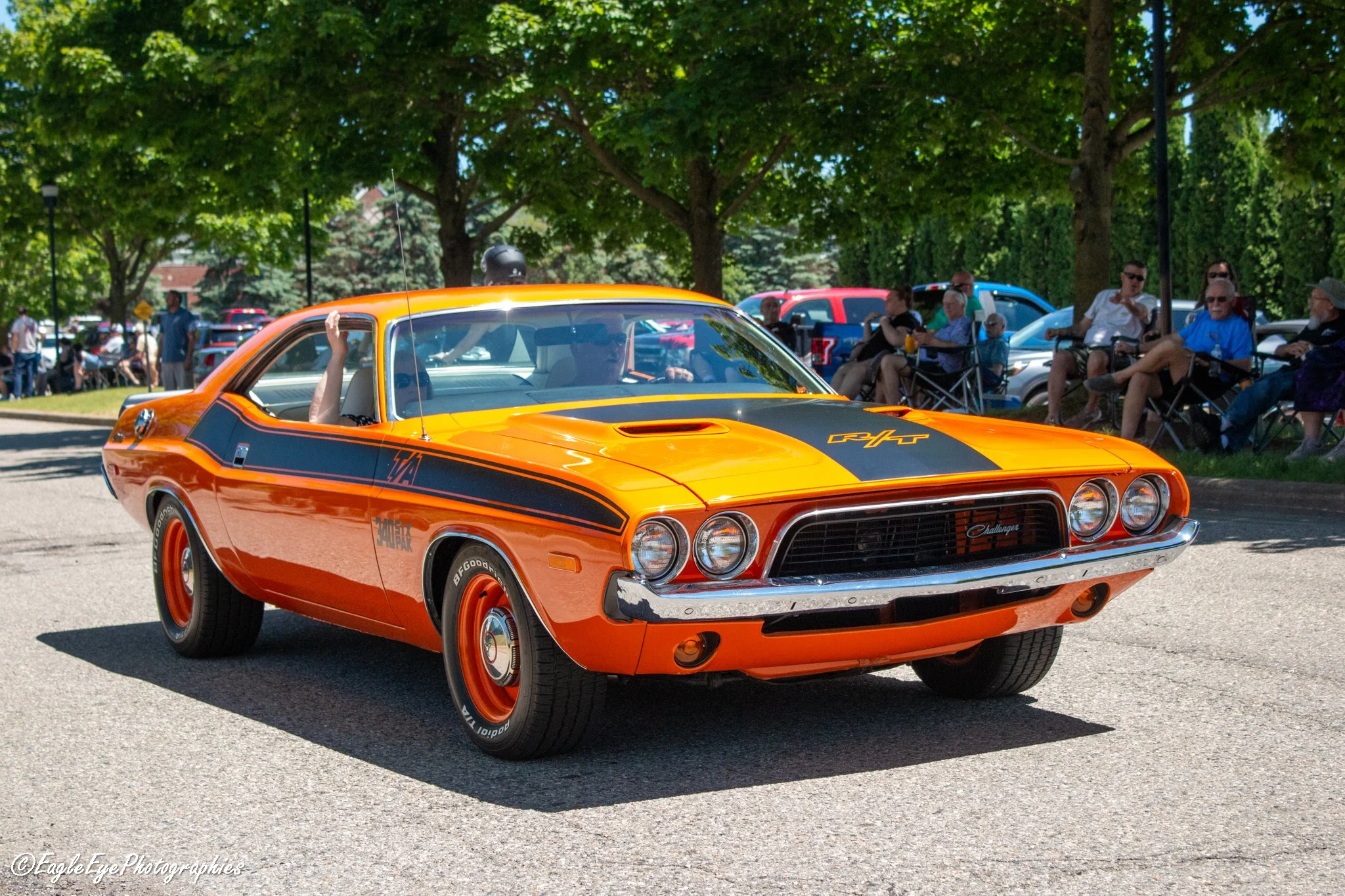 An orange classic Dodge Challenger muscle car with black racing stripes driving on a street during a parade or car show, with spectators sitting on chairs under trees in the background.