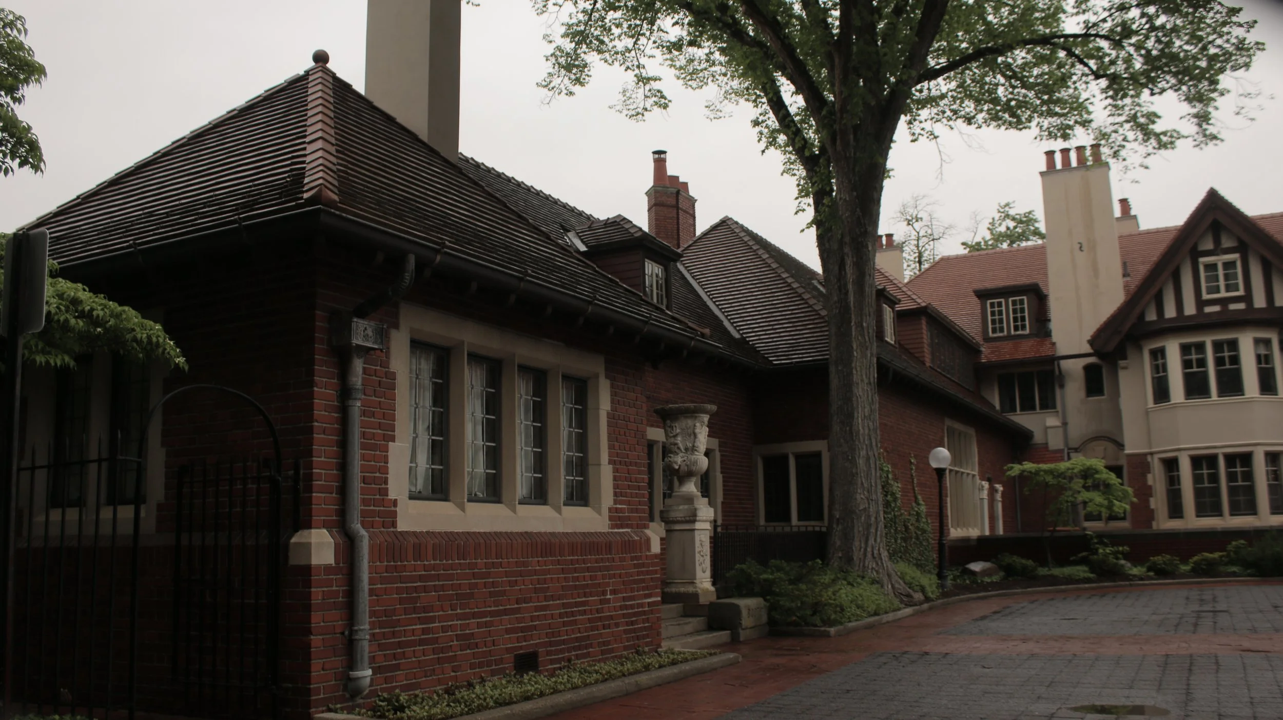 A neighborhood street with large, historic brick houses and lush trees. The houses have gabled roofs, multiple chimneys, and decorative architectural details.