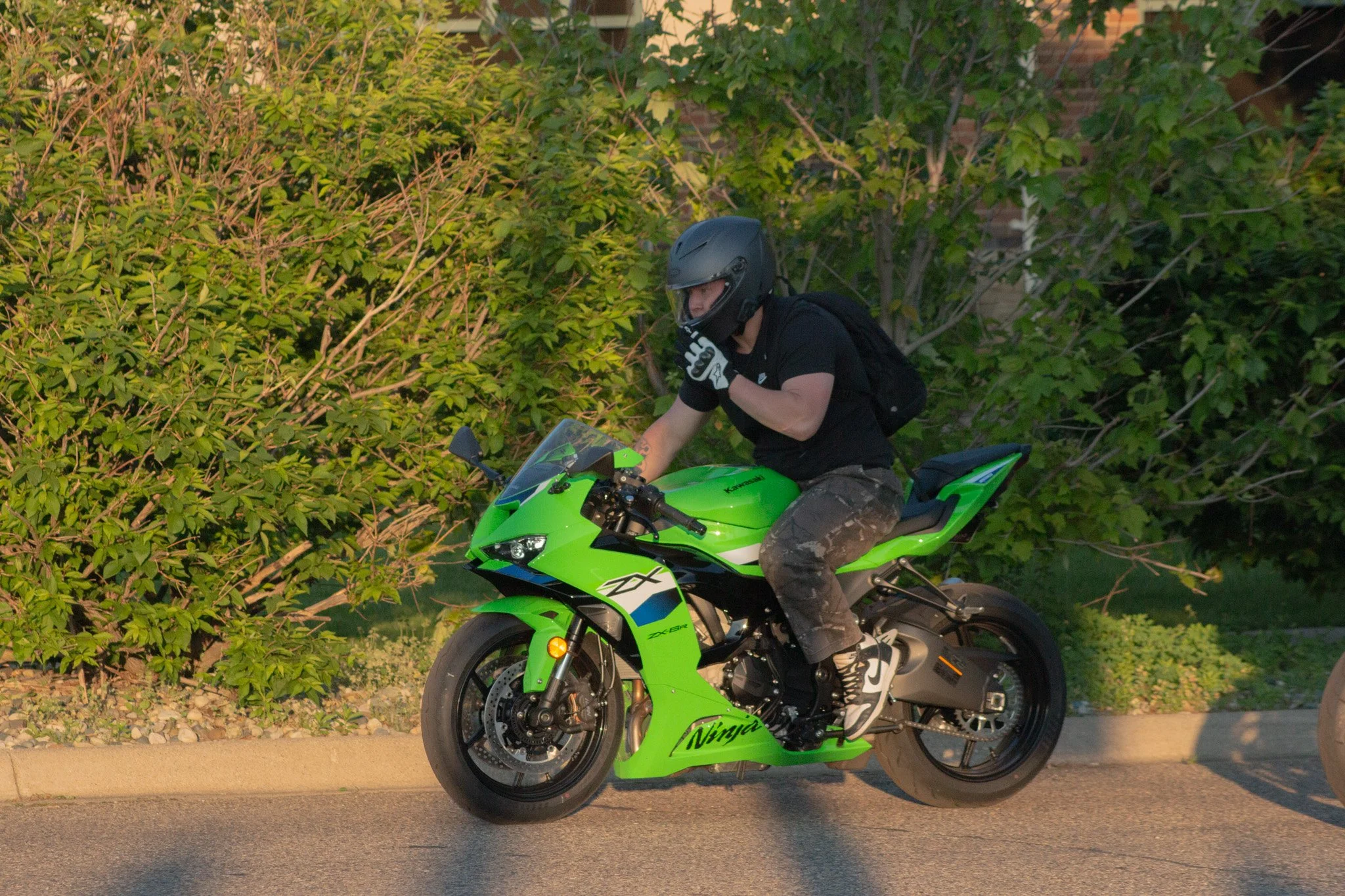 A person wearing a black helmet and black shirt riding a bright green Kawasaki Ninja sport motorcycle on a city street beside a tree with green foliage.