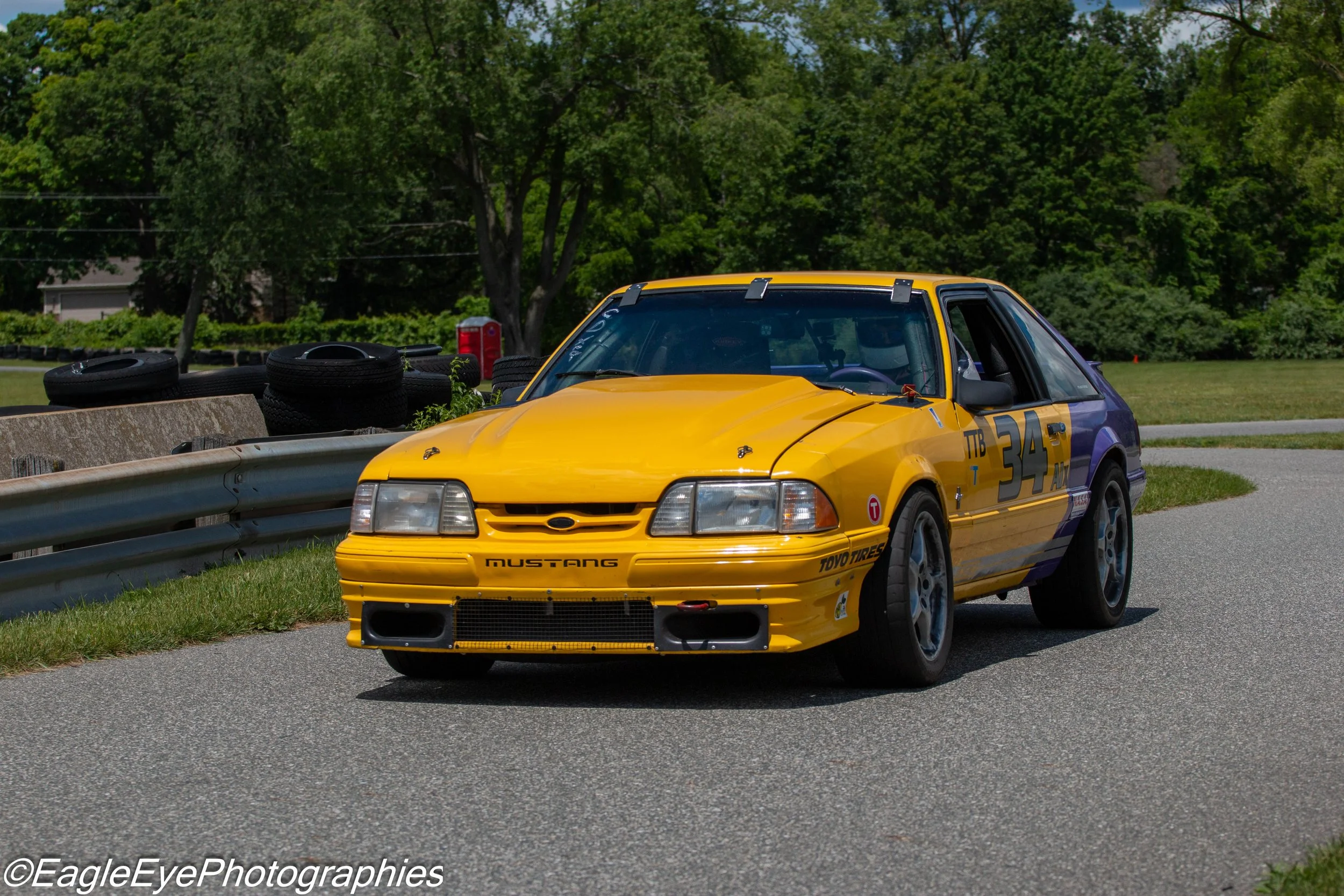 A yellow race car with racing numbers parked on the side of a road next to a guardrail, with black tires stacked on a concrete barrier and green trees in the background.