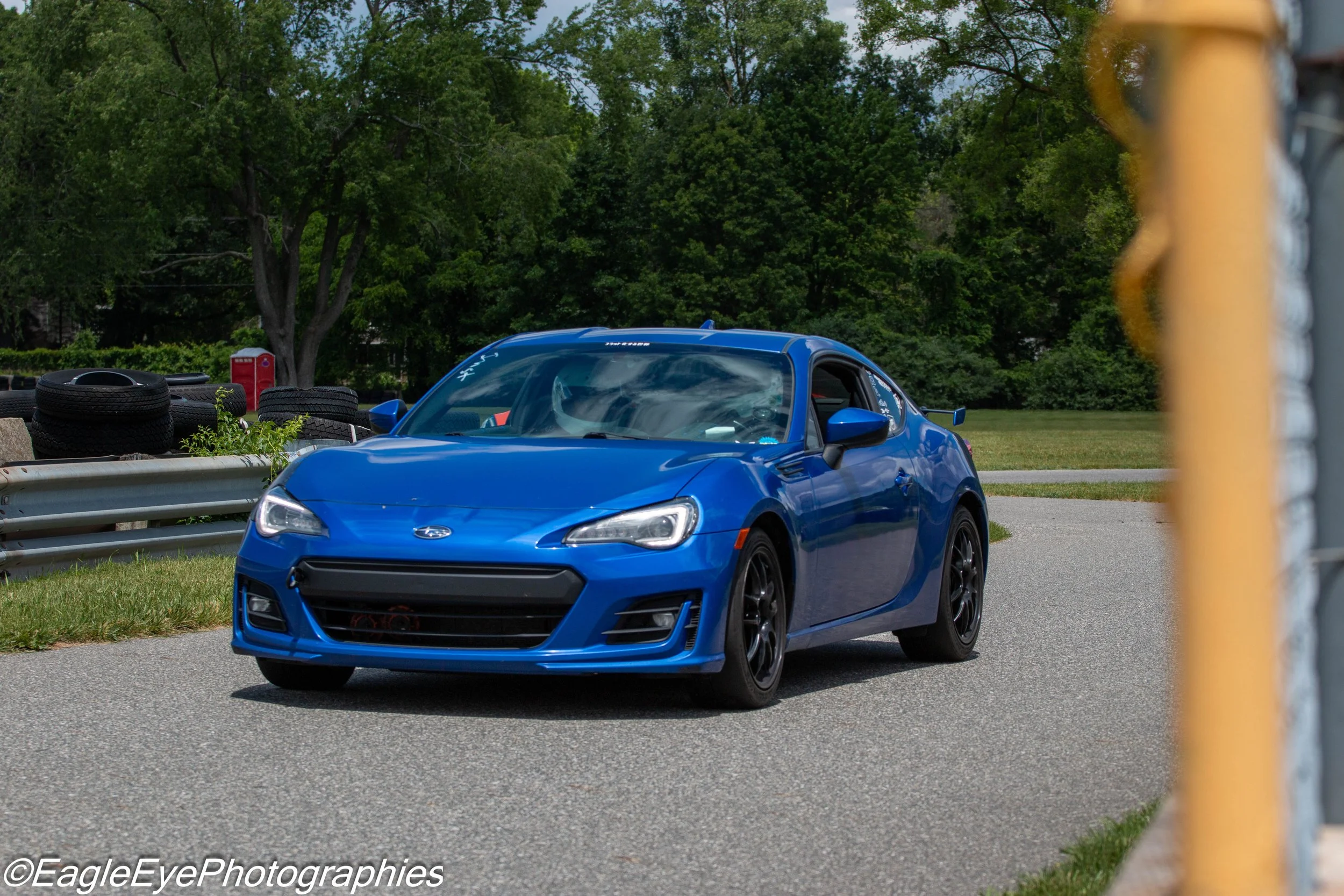 A blue sports car parked on a road beside a grassy area with tire stacks and a red mailbox, trees in the background, and a yellow fence post partially visible on the right.