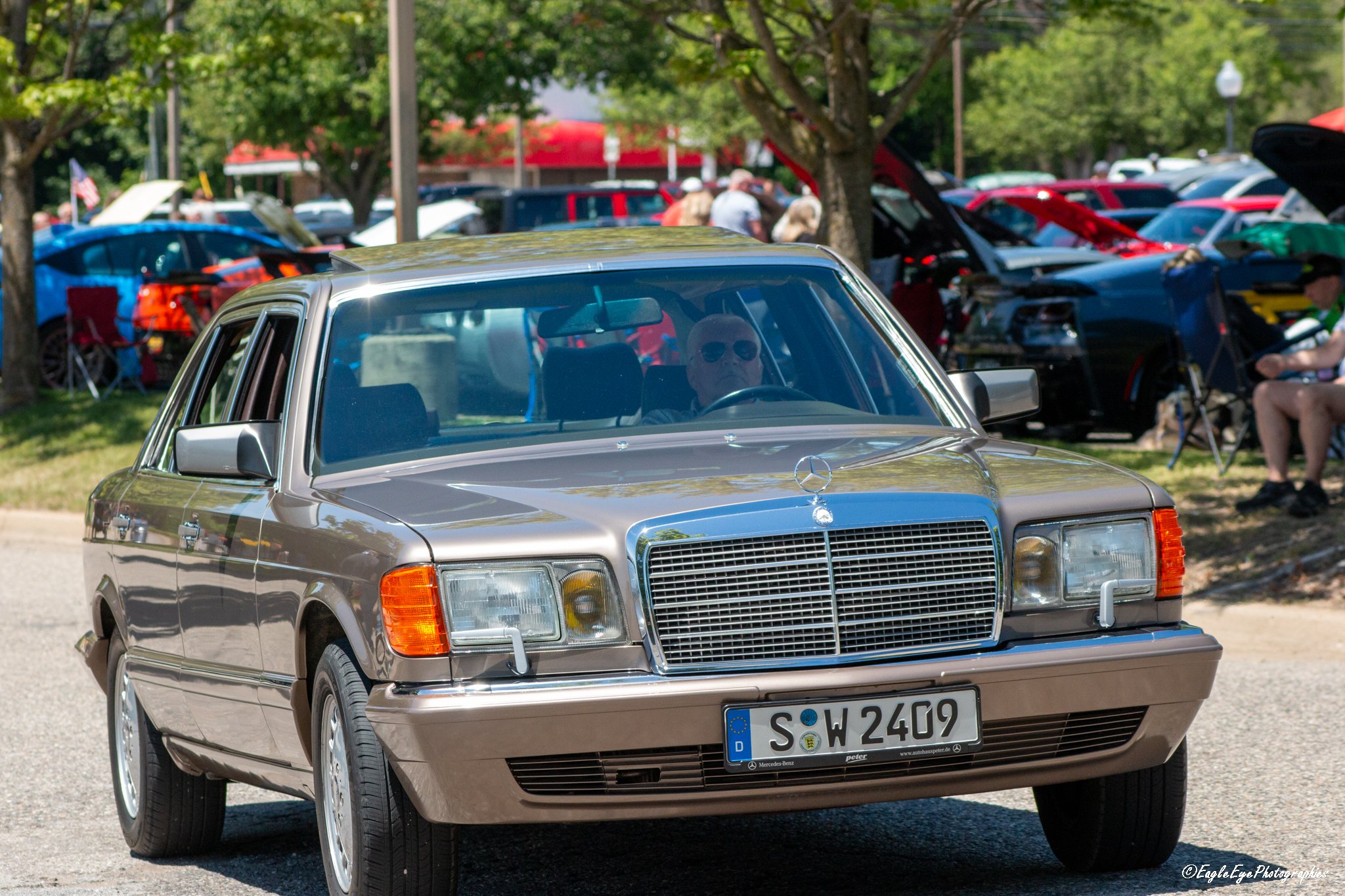 A vintage silver Mercedes-Benz car at a car show, with a woman wearing sunglasses sitting behind the wheel. In the background, other cars with open hoods and people are visible under trees on a sunny day.