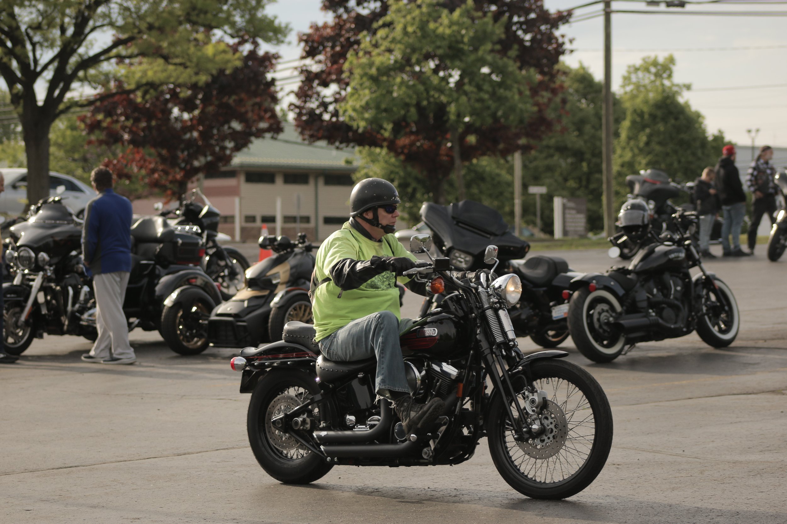 A man wearing a helmet and sunglasses riding a black motorcycle in a parking lot filled with other motorcycles. Several people are standing and walking nearby, with trees and a building in the background.