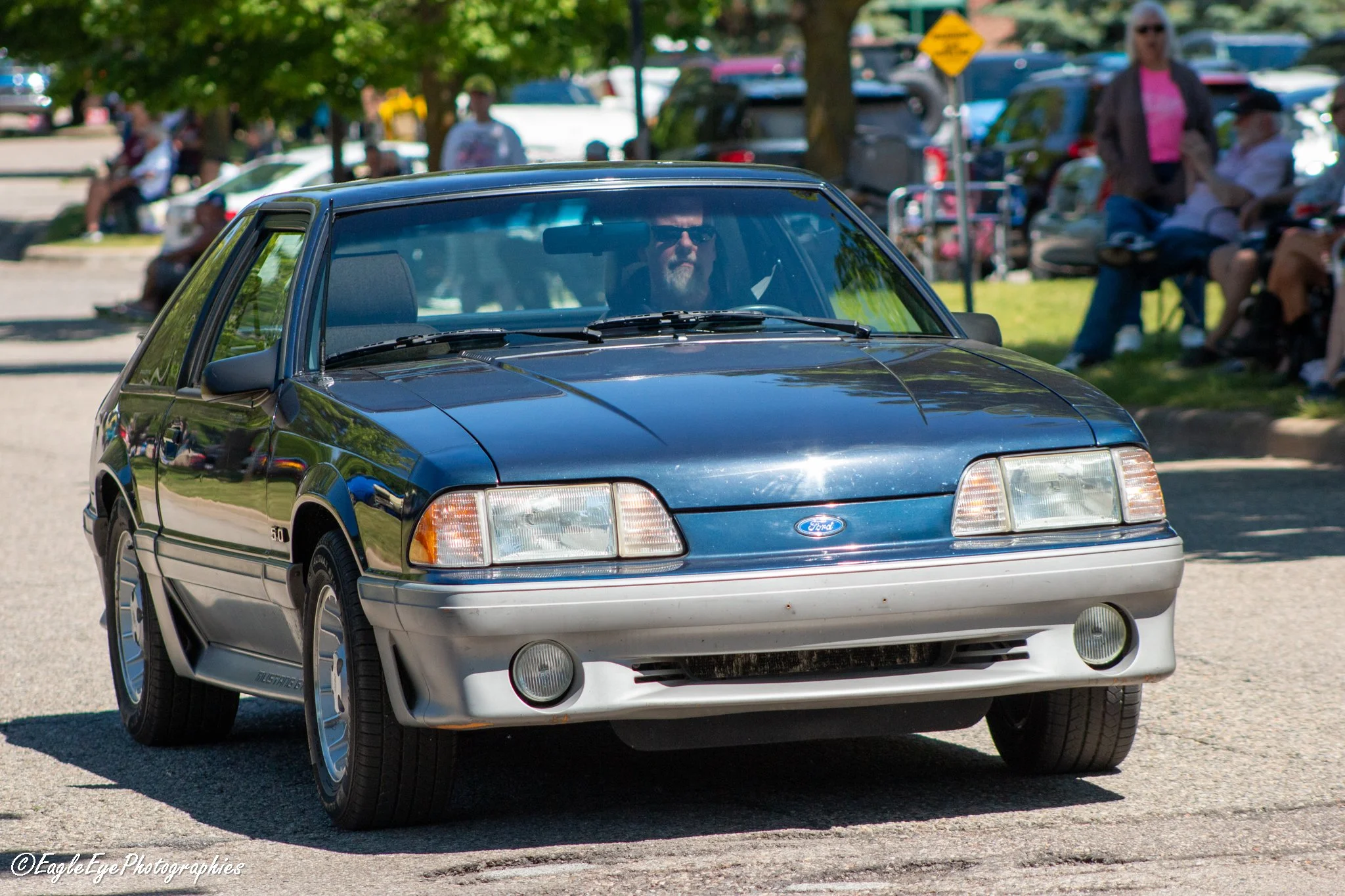 A vintage blue and gray Ford Mustang car parked on a street with people sitting and standing in the background during daytime.