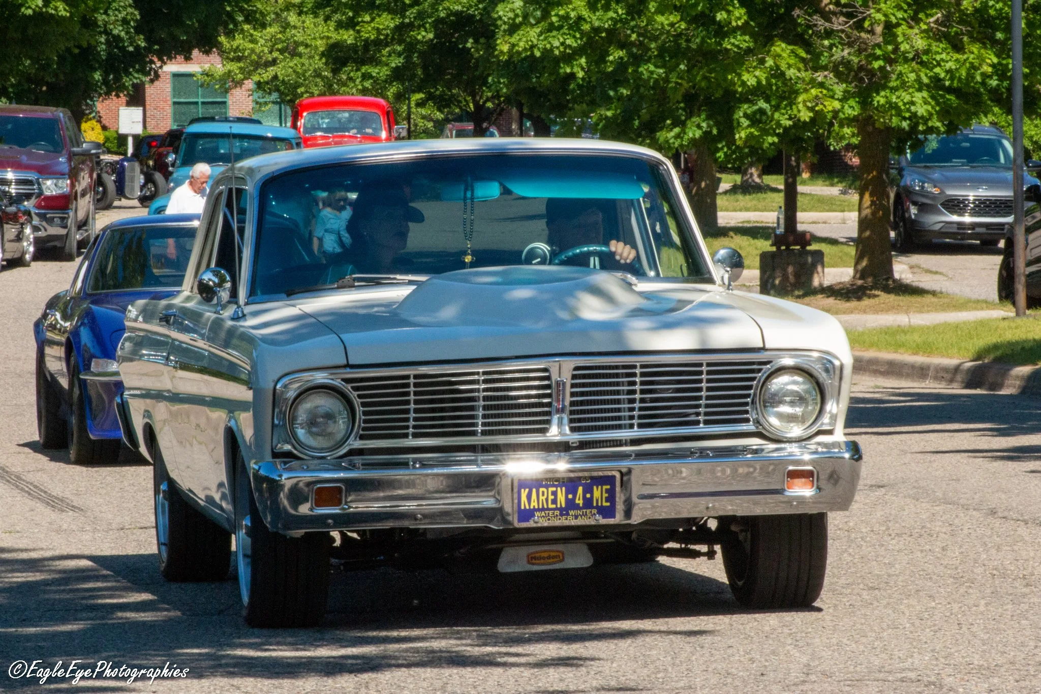 A classic silver car with a custom license plate reading 'KAREN-4-ME' is driving down a street lined with parked cars and green trees. The driver is reaching with their right hand, and there are people standing near a red vintage truck in the backgro