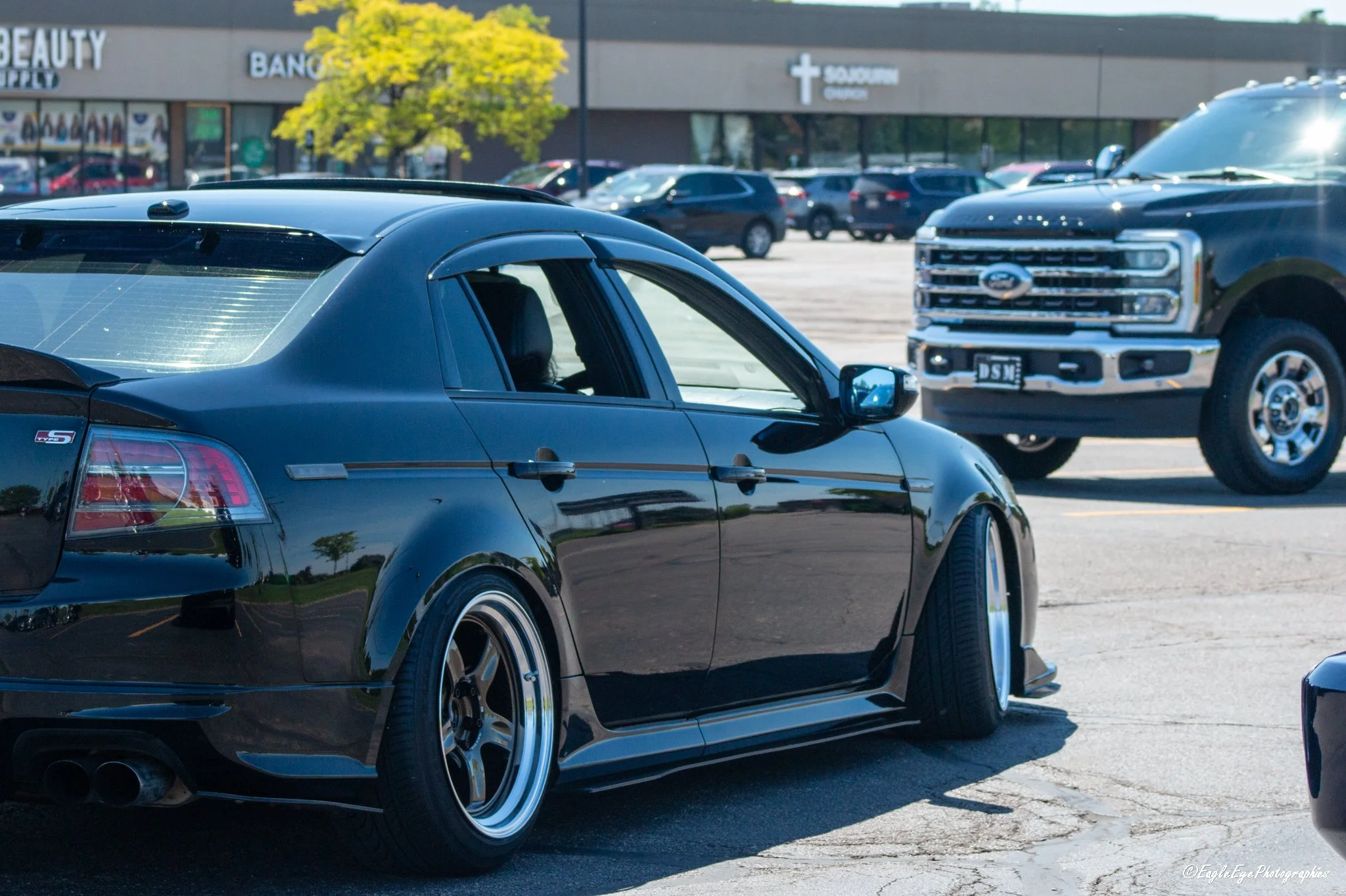 A black car with custom rims and a lowered suspension parked in a parking lot. In the background, there are other vehicles, a tree with green leaves, and a shopping center with signage for beauty supplies, a bank, and a Southern Oregon pharmacy.