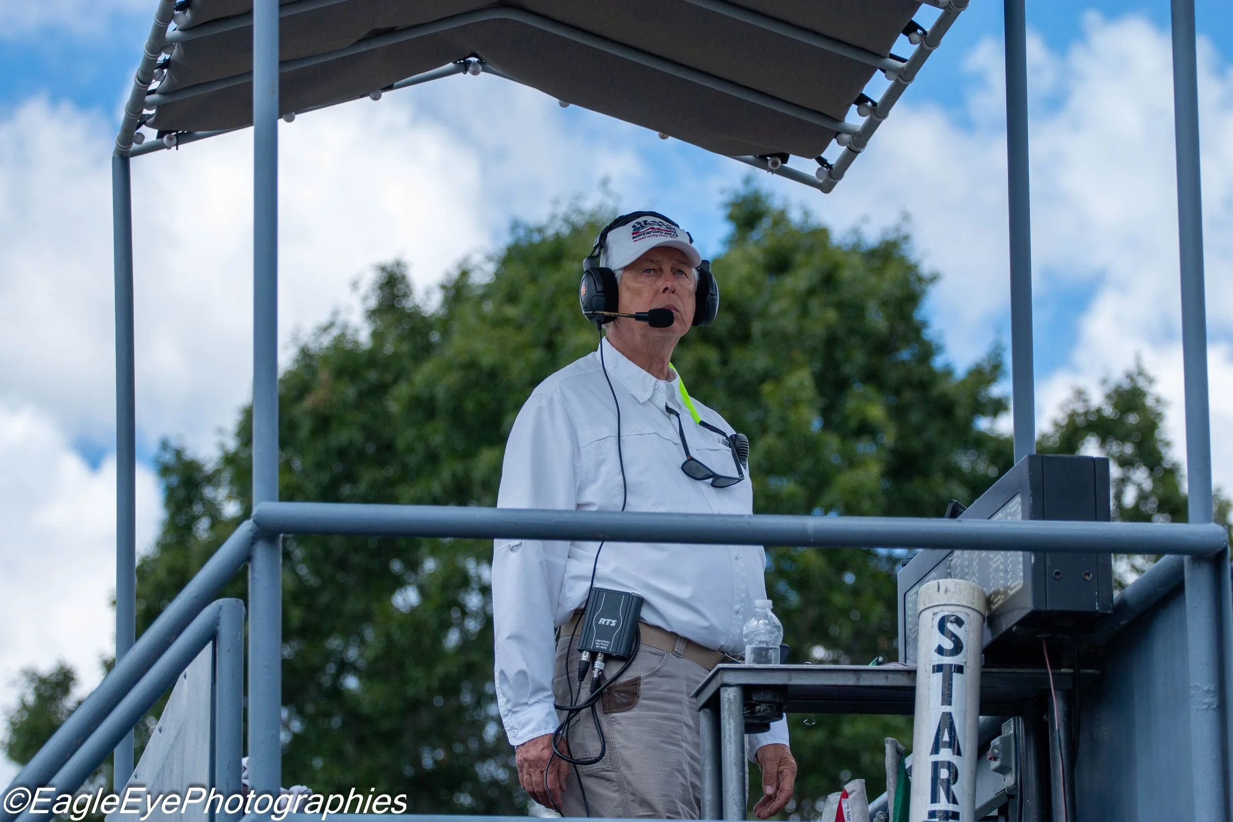 An older man operating equipment on a raised platform outdoors during daytime. He is wearing a white cap, a white long-sleeve shirt, and a headset with a microphone. The background shows green trees and a partly cloudy sky.