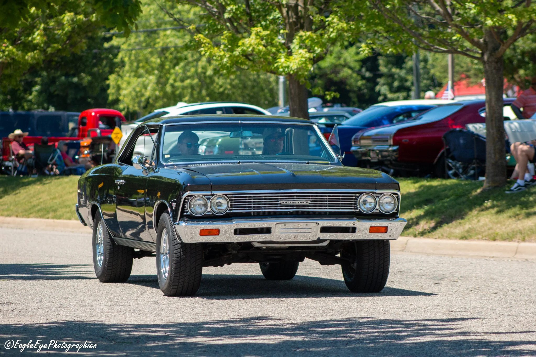 A black vintage Chevrolet Corvette car driving on a street with green trees and parked cars and people in the background during daytime.