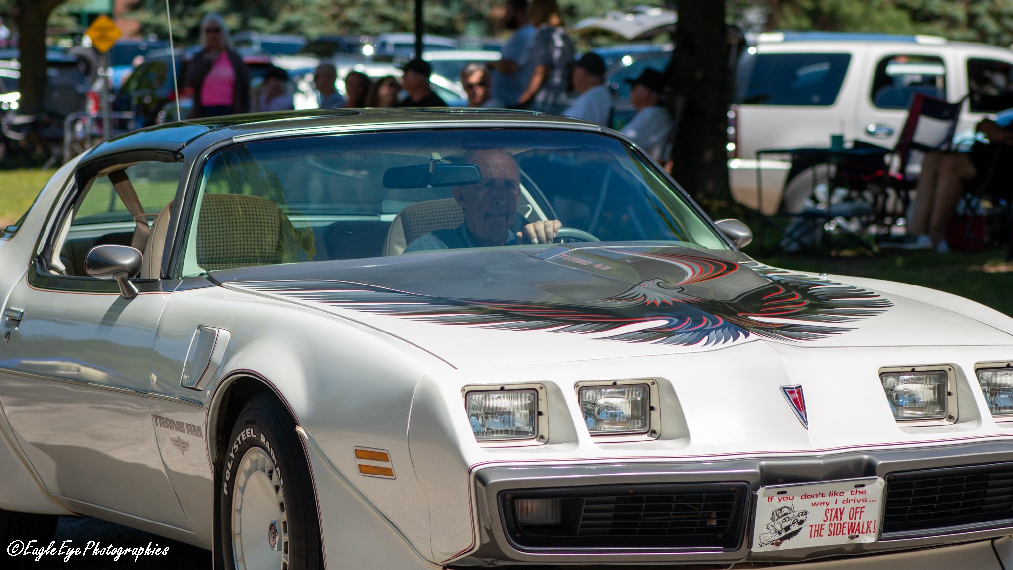 A white Pontiac Trans Am with a black and red eagle graphic on the hood is parked at an outdoor car show. People are gathered in the background, and there are trees and other vehicles around.
