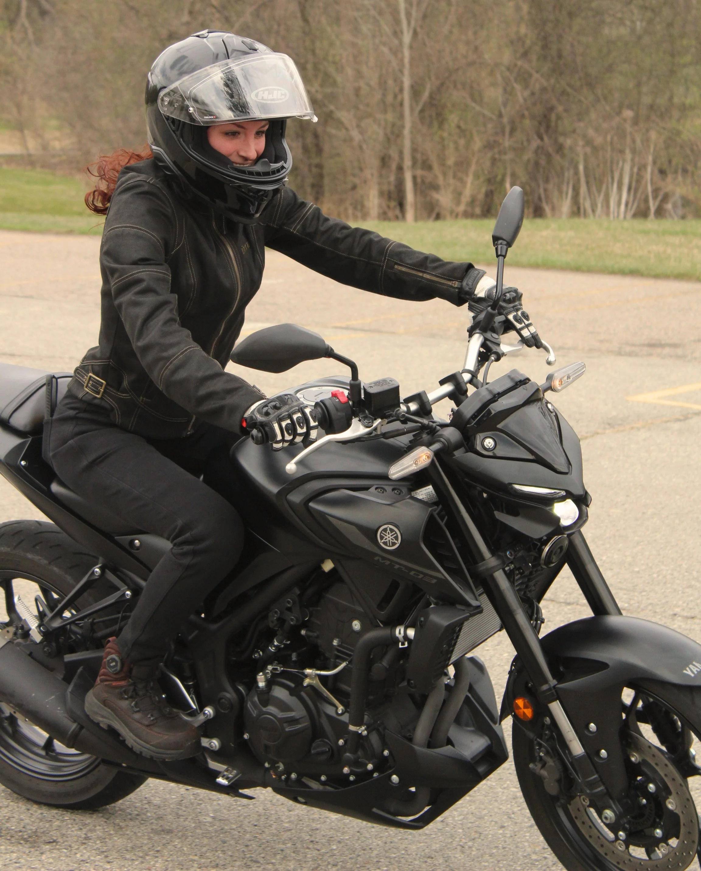 A woman riding a black Yamaha motorcycle, wearing a black helmet and black jacket, on a paved road with trees in the background.