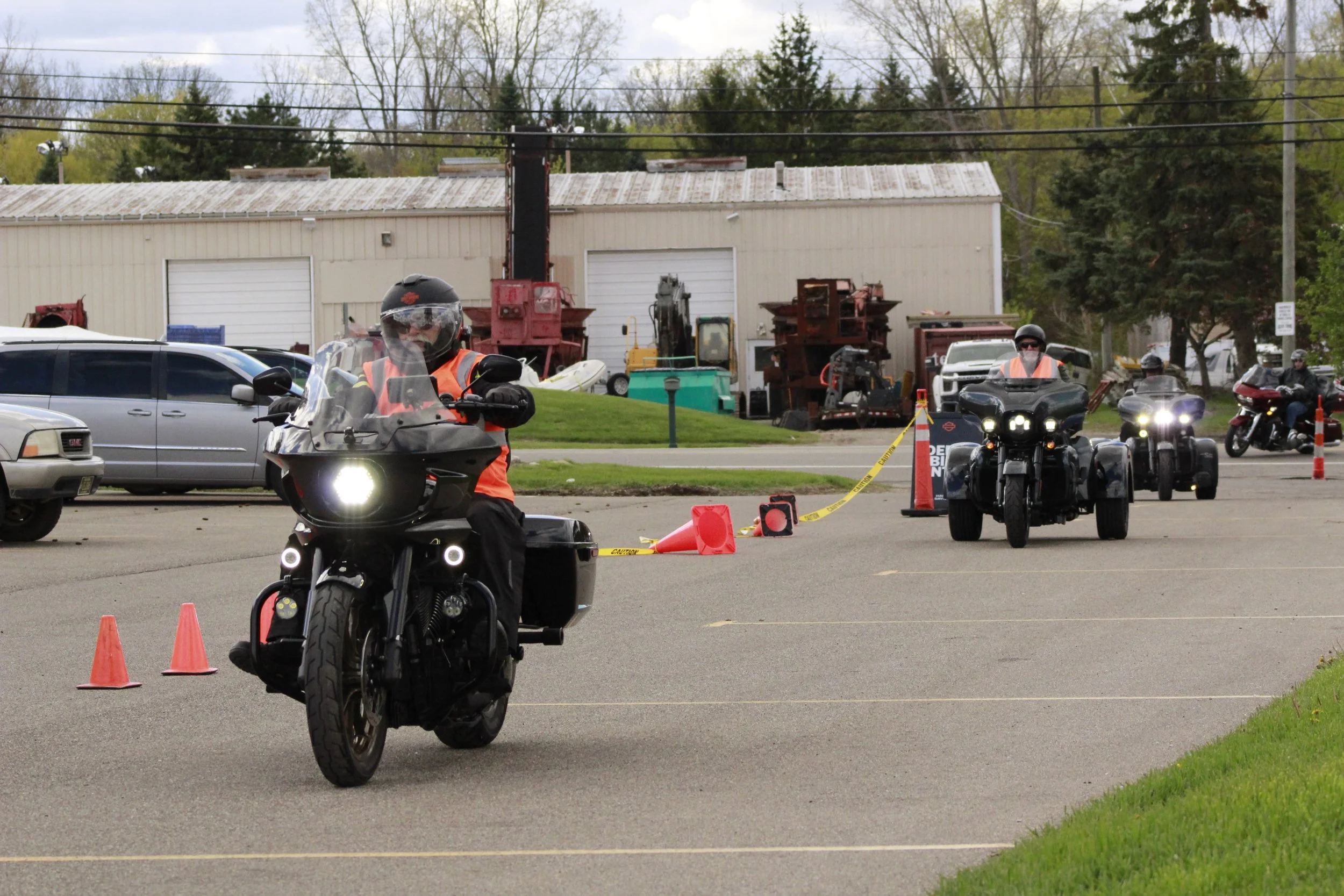 Motorcyclists riding in a line through a parking lot with orange cones, with a building and trees in the background.