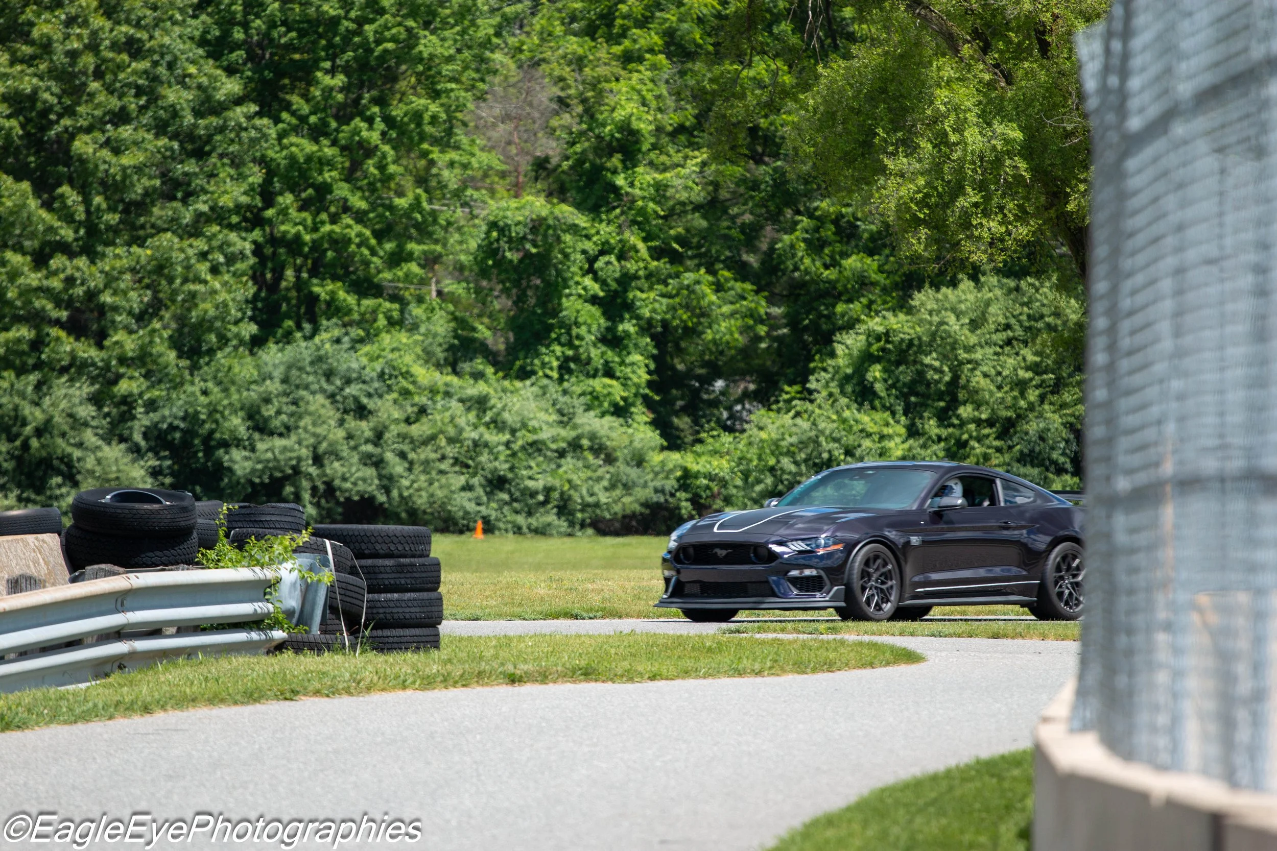 A black sports car driving on a race track surrounded by green trees and tires stacked along the side of the track.
